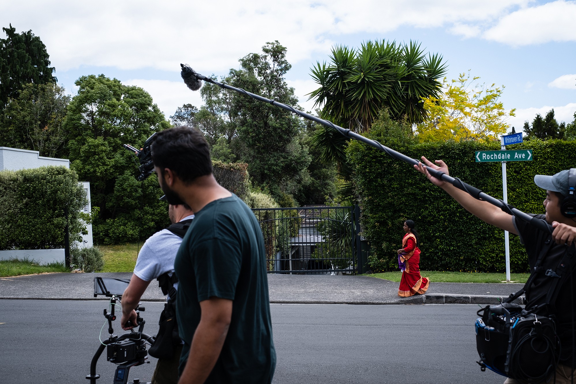 A woman in a bright red sari walks along the footpath on a suburban street while three men standing in the foreground point their video and sound equipment in her direction.