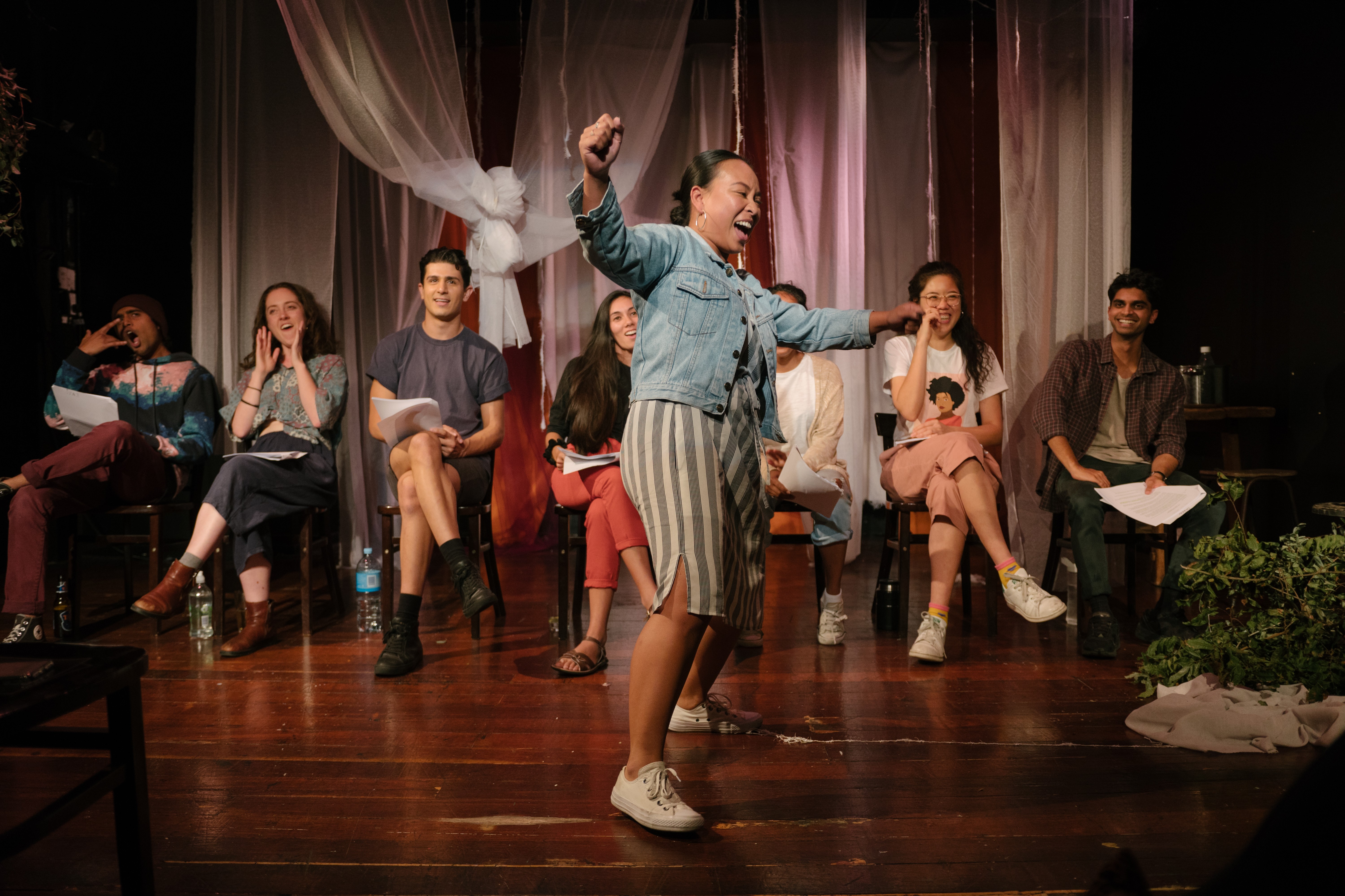 Seven actors sit on a line of chairs with scripts on their lap. A woman stands in front of them dancing and smiling