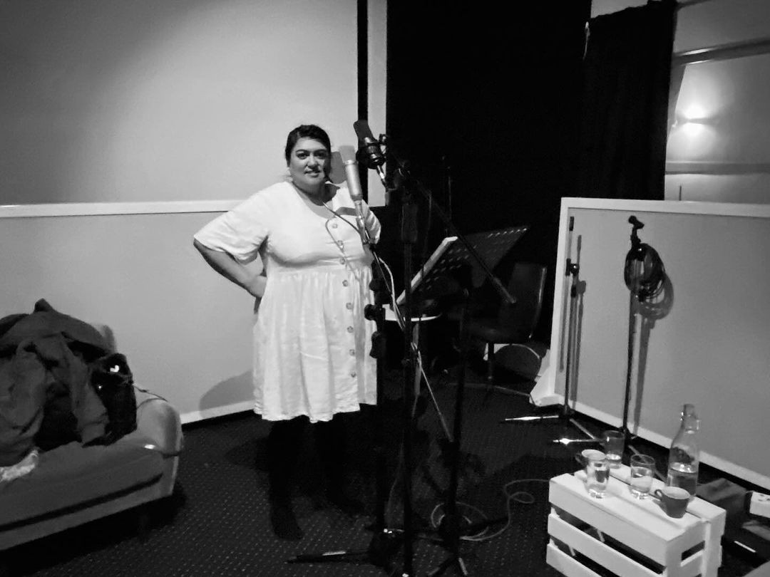 Black and white image of a woman in a white dress with her hands on her hips surrounded by a microphone, and a stand holding her script