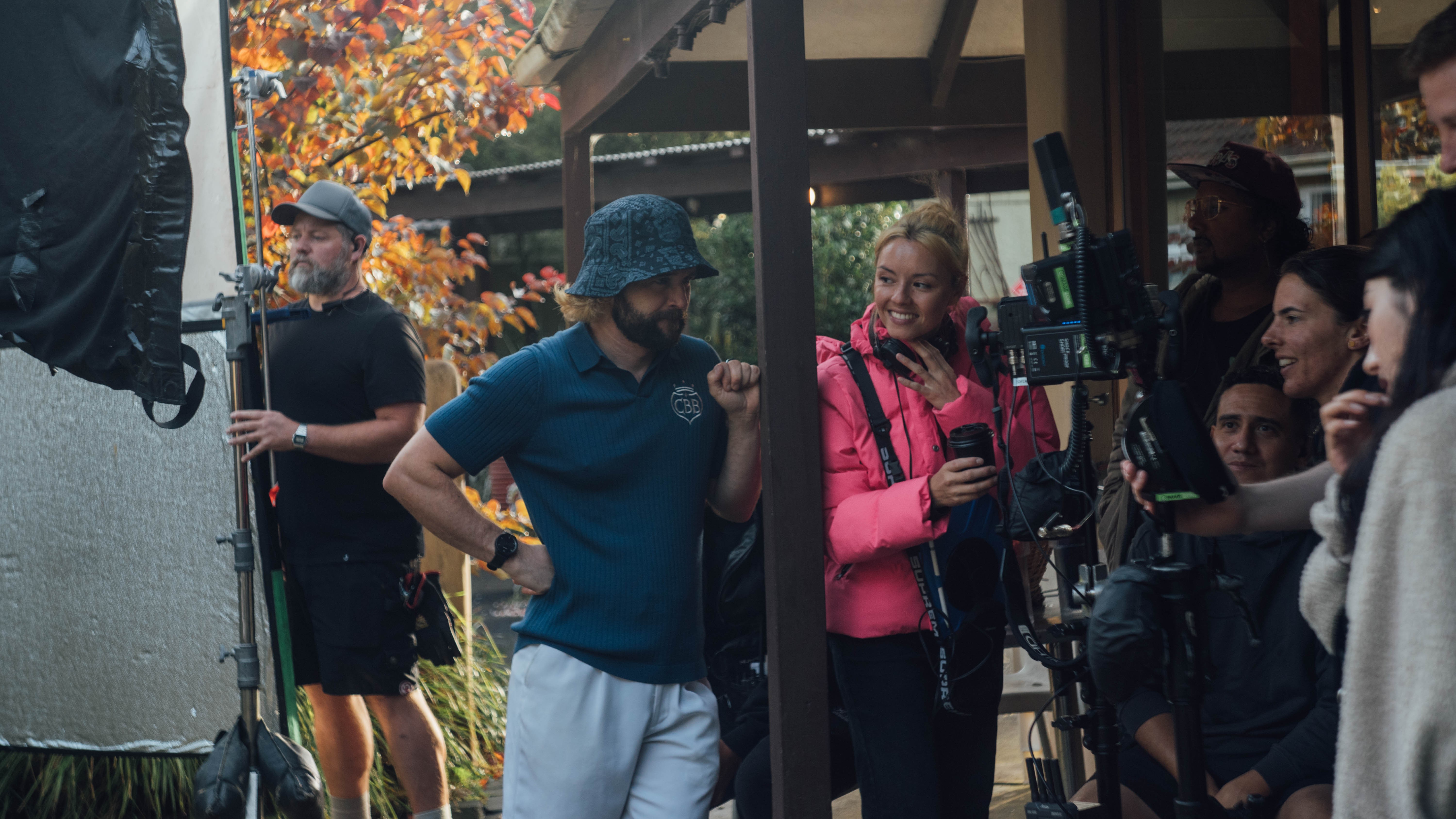 Two people standing behind a camera. A woman in a pink puffer jacket and a man in a denim bucket hat