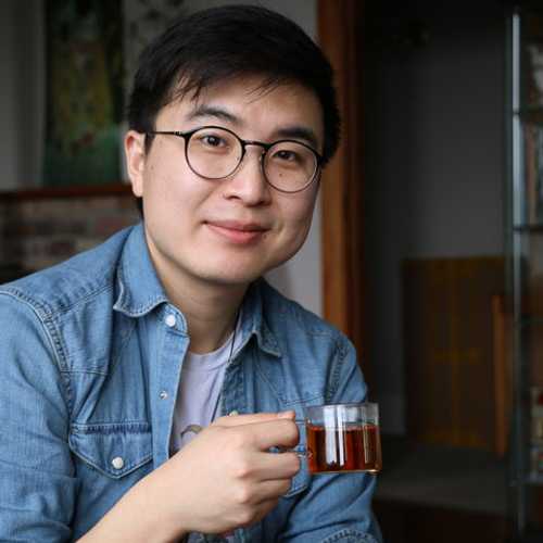A photo of a person holding a cup of coffee behind shelves of books and various figurines.