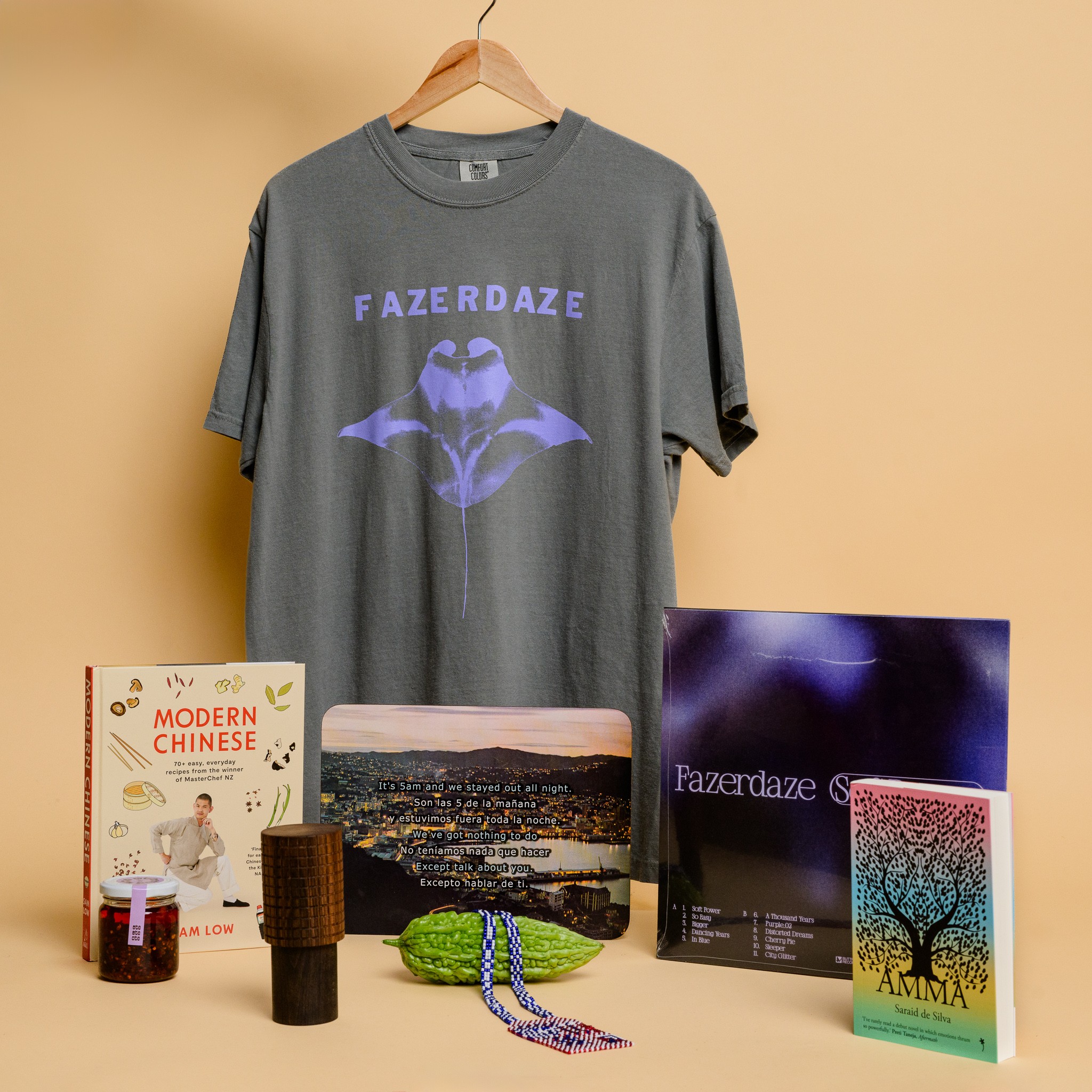 A grey t-shirt with a lilac stingray printed on it with the text 'Fazerdaze', a cookbook and chilli oil, a grinder (pepper mill), a beaded necklace resting on a bitter gourd, a placemat featuring city lights and text overlaid, an album and a book