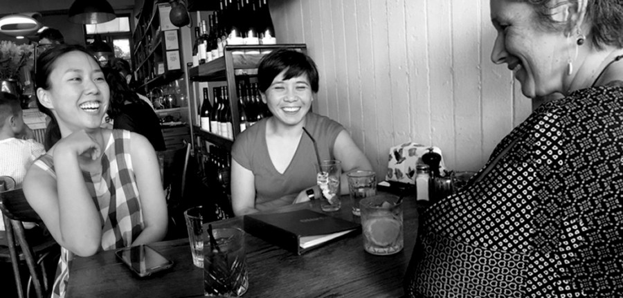 Black and white photo of three women laughing around a cafe table. 