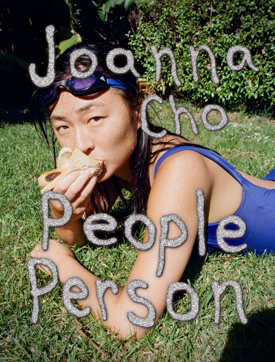 Photo of an Asian woman lying on the grass in a blue swimsuit wearing goggles and eating a sandwich.