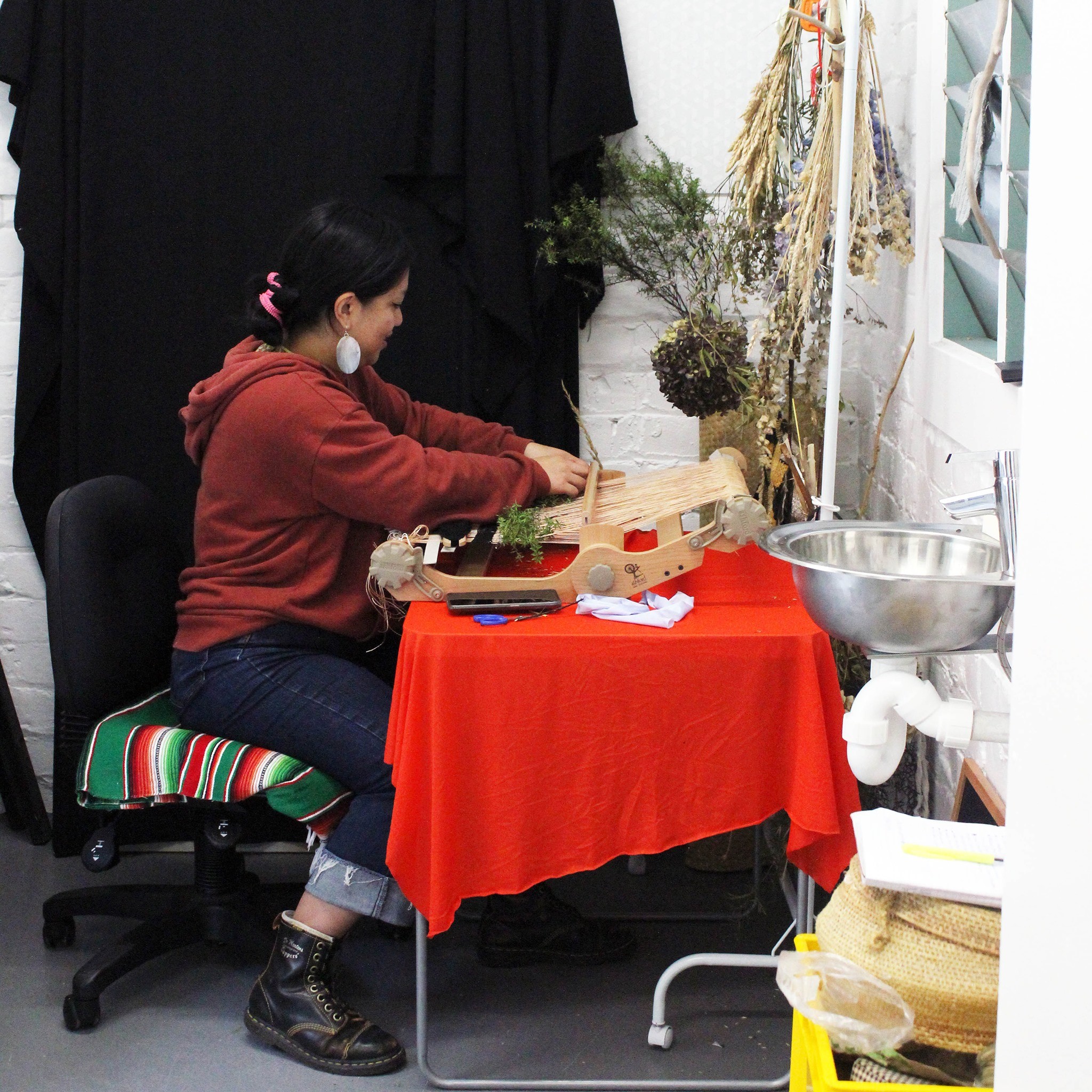 A person of Filipino descent in a maroon jumper weaving on a loom placed on a red table