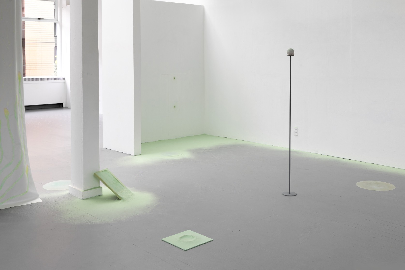 A plain room with white walls and a grey floor with small objects scattered around spraypainted pastel green, the green spilling onto the floor surrounding.