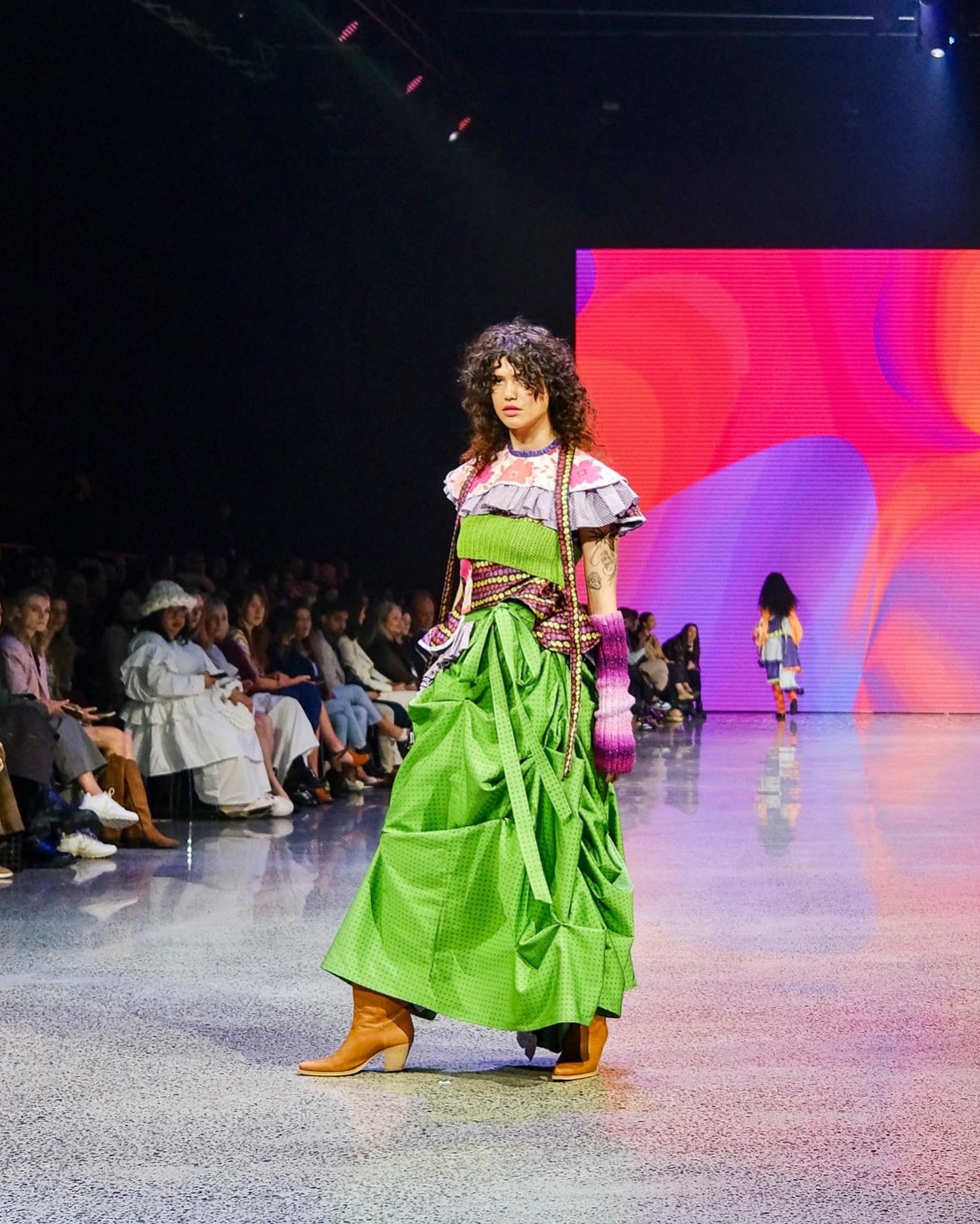 A person in a colourful and stylishly mis-matched avant-guarde outfit walking a fashion show with seated people watching them from the sides