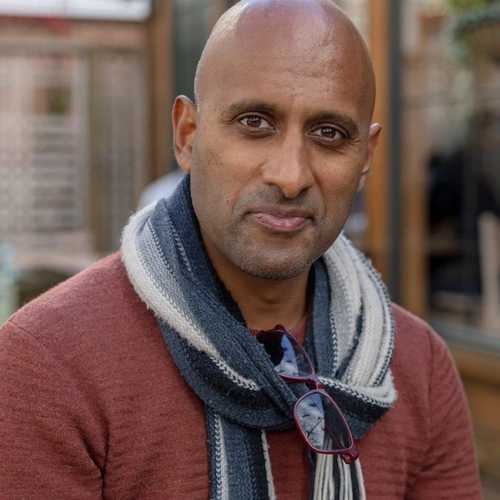 Brannavan Gnanalingam is wearing a red sweater with a striped scarf. He is bald and smiling for the camera..