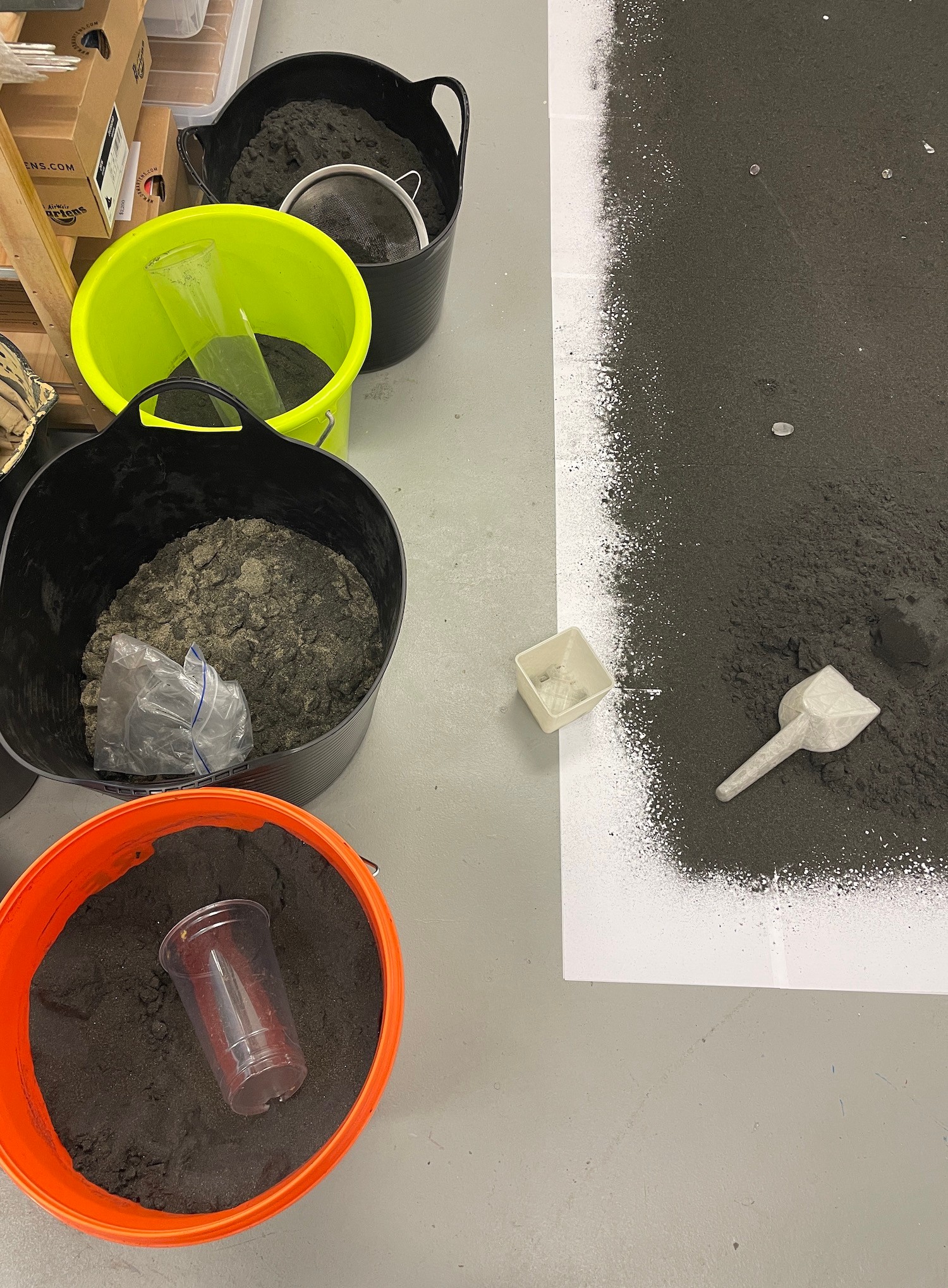 Plastic buckets and containers containing black sand next to some arranged on the floor. 