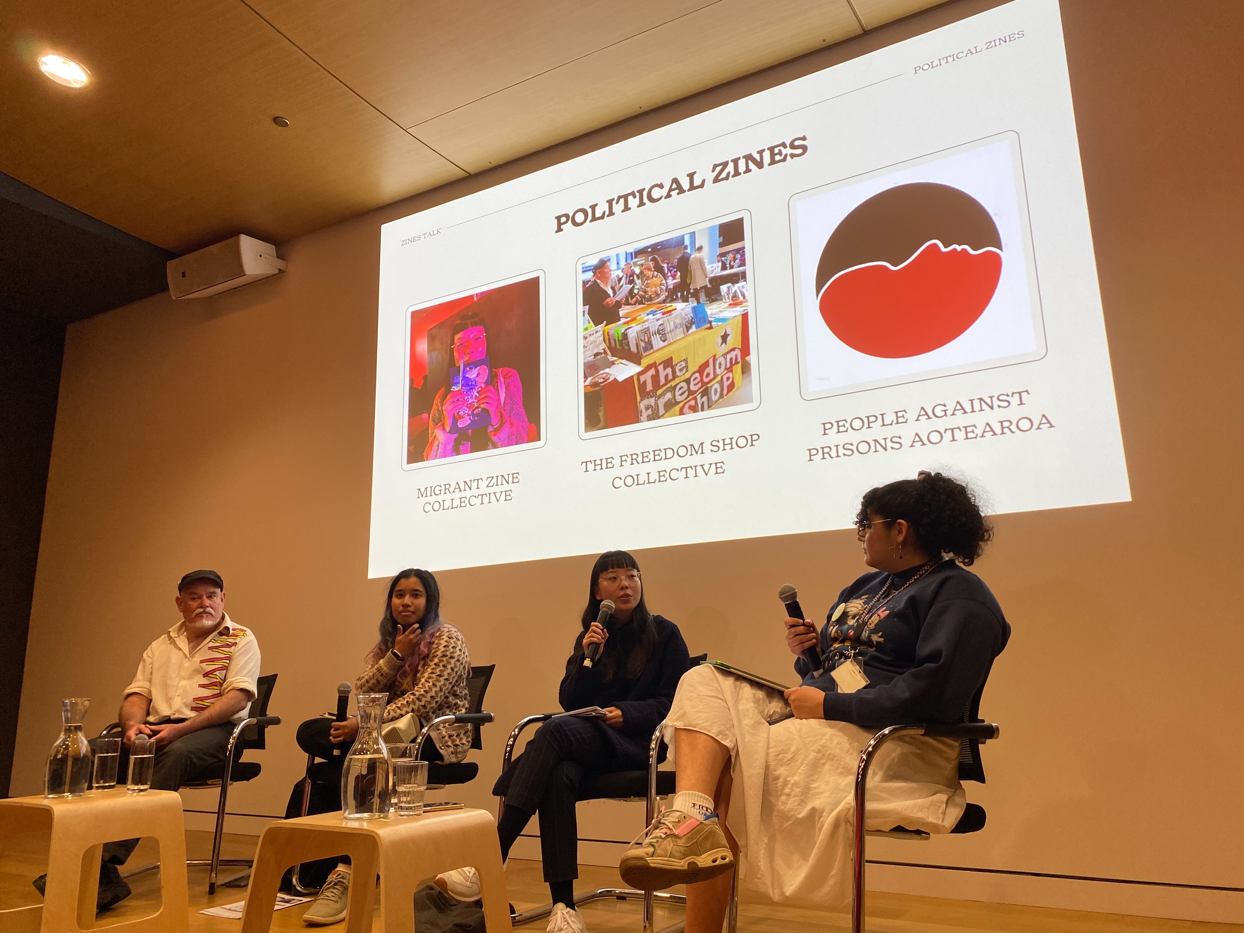 Four people seated with microphones on a stage with a projection behind displaying the words "Political Zines".