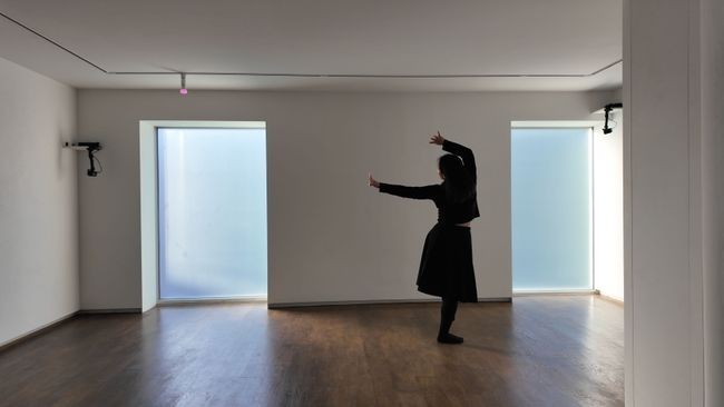 A woman in a long-sleeved black top and long black skirt dances in an empty space with wooden floors, light diffusing in through two floor-to-ceiling windows.