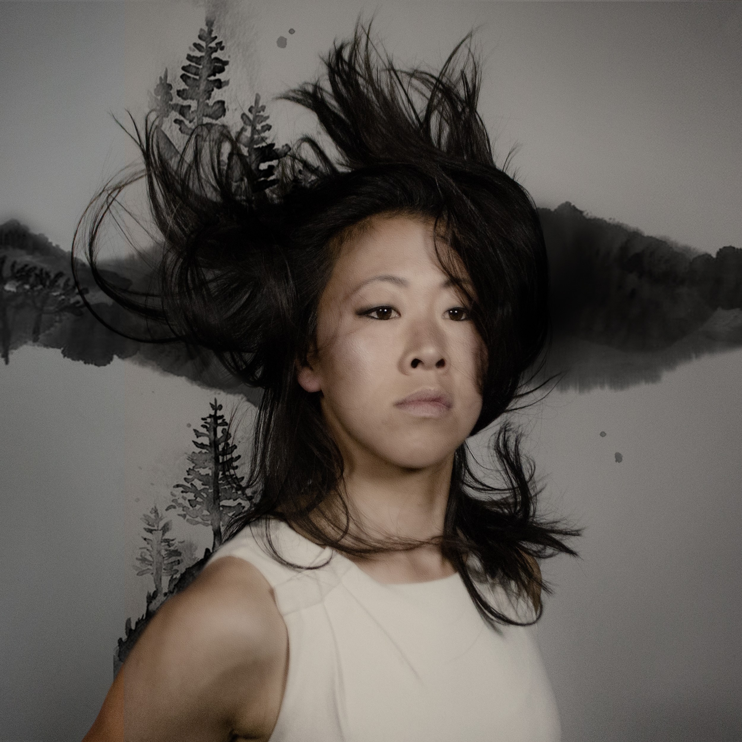 A woman with her black hair flying in the air against a grey background with shadows of trees and leaves featuring in the back wall.