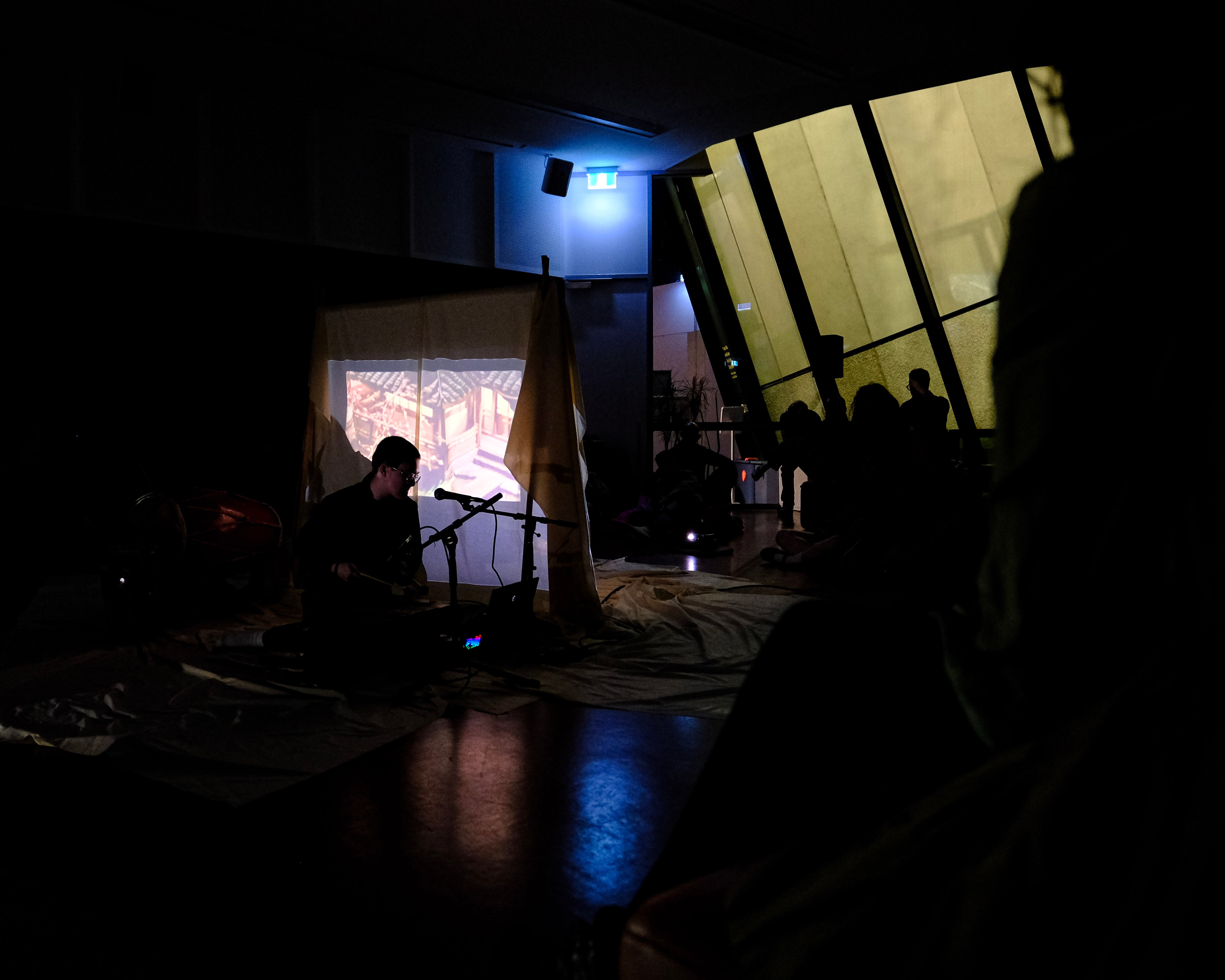 A darkened room with a few lights and projections cast. The silhouette of the artist and a microphone is visible from a distance.