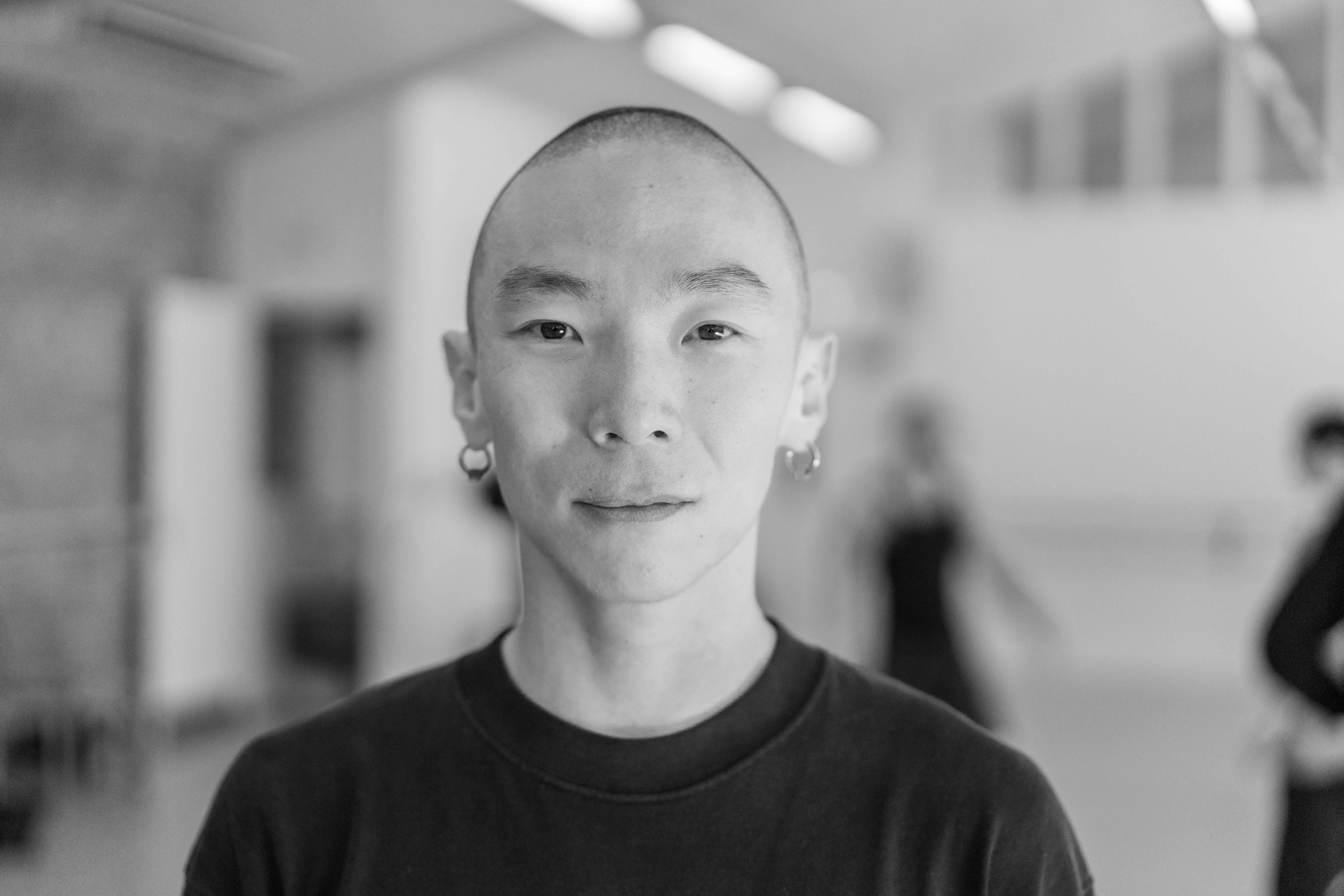 A black and-white photo of a man with a shaved head and earrings gazing directly at the camera