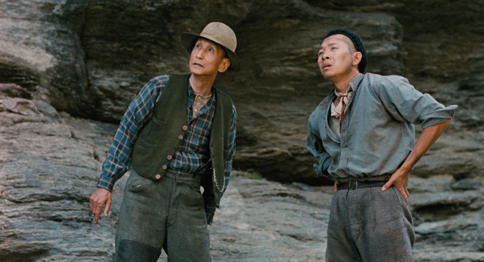 In a scene from “Illustrious Energy”, two Chinese men stand with their backs against a rocky cliff face, looking into the distance