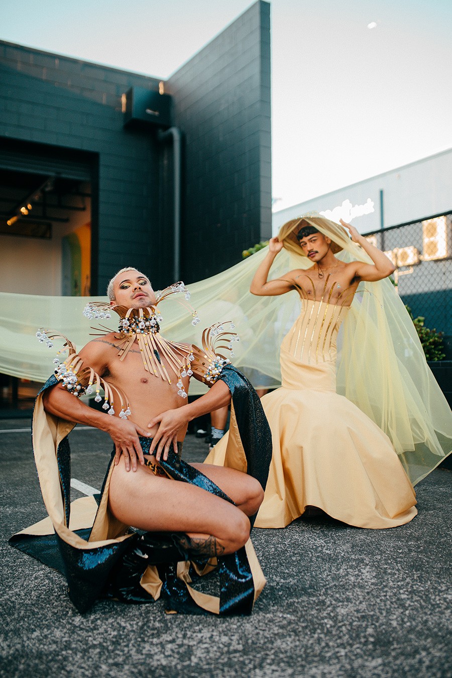 Two gender-bending models: one crouching low wearing a sparkly black skimpy outfit with huge beads, the other wearing a gown with the chest exposed and a wide brimmed hat attached with flowy yellow translucent fabric