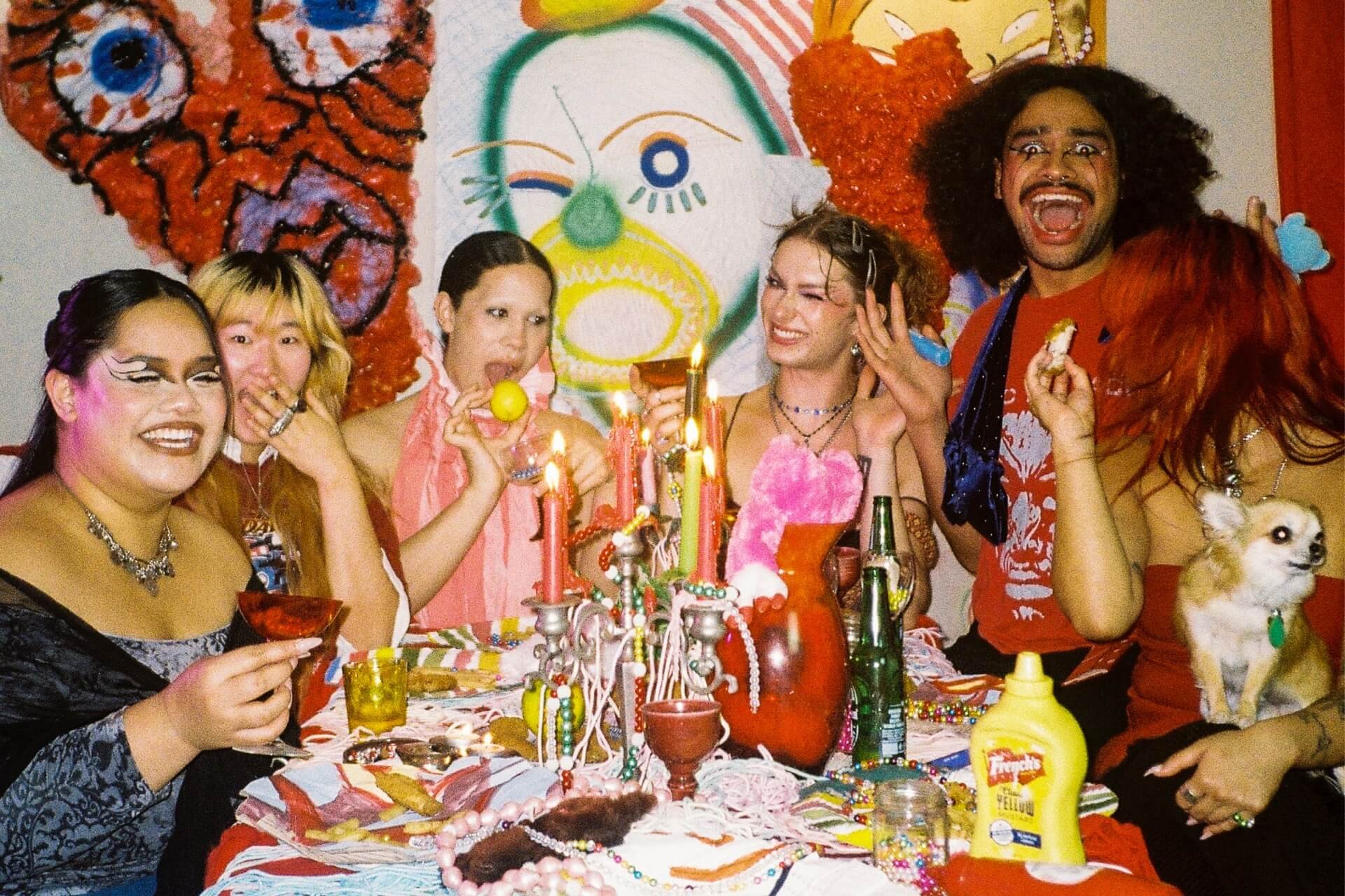 6 fashionable dressed people doing various camp poses seated in front of a festive and messy table featuring various finger food and props