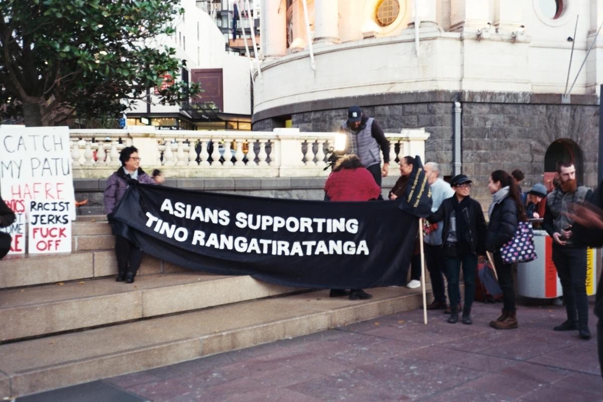 A group of people holding a large black banner saying 'Asians Supporting Tino Rangatiratanga'