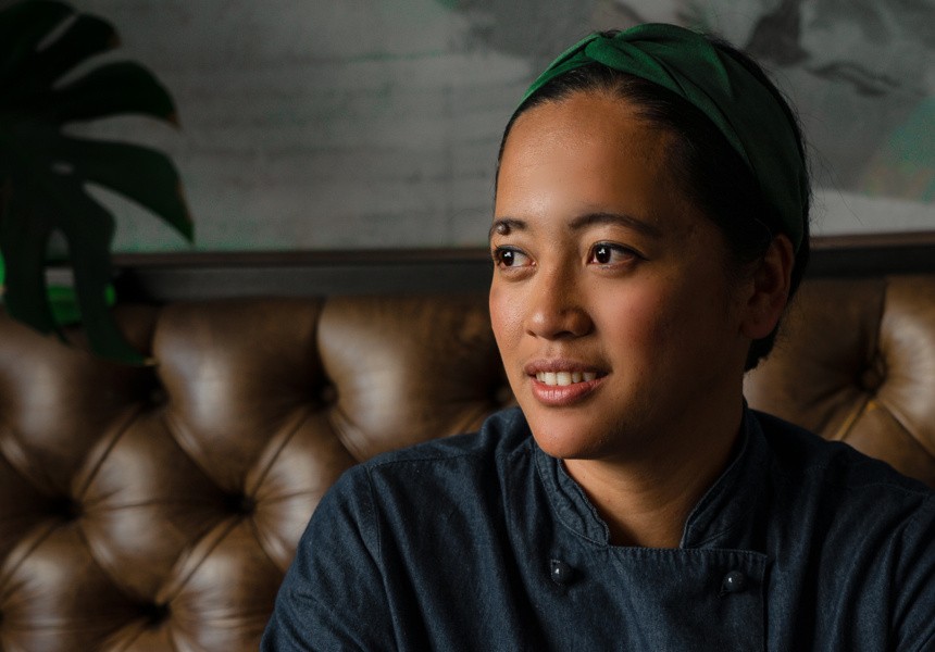 Dramatic lighting of a smiling person of Filipino descent wearing a blue chef's uniform and sitting on a brown sofa