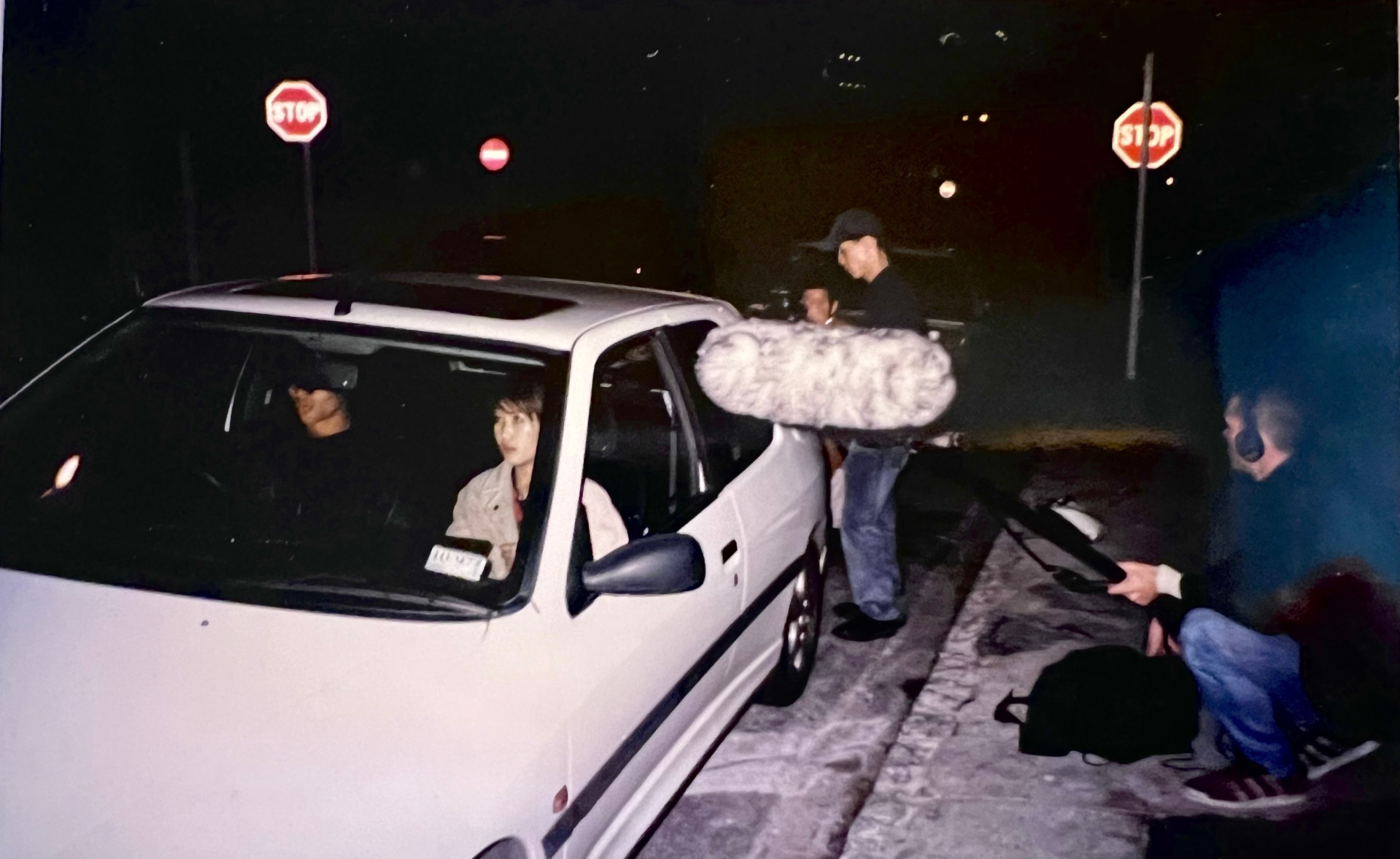 At night, a director and his film crew prepare to shoot two actors inside a white hatchback car.