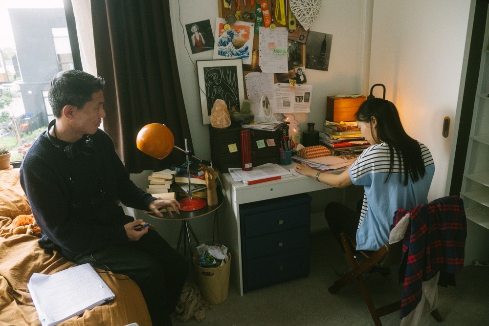 On the set of a film shoot, a man sits on a bed across from an actor sitting at a desk.