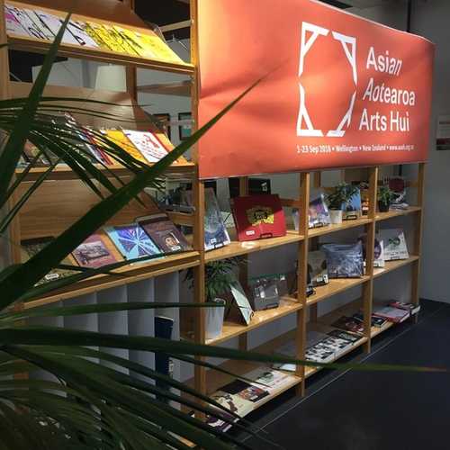 Shelving in a library with a zine display and Asian Aotearoa Arts Hui banner.