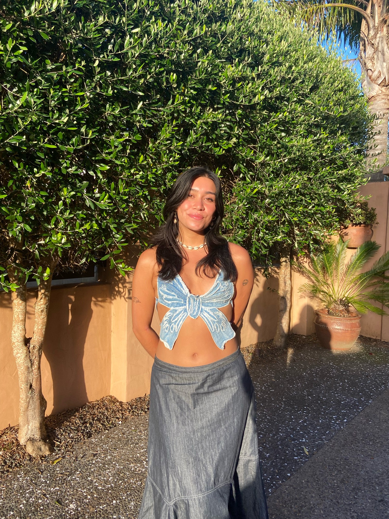 Person in a blue ribbon top and long grey skirt wearing a shell necklace being photographed during the golden hour