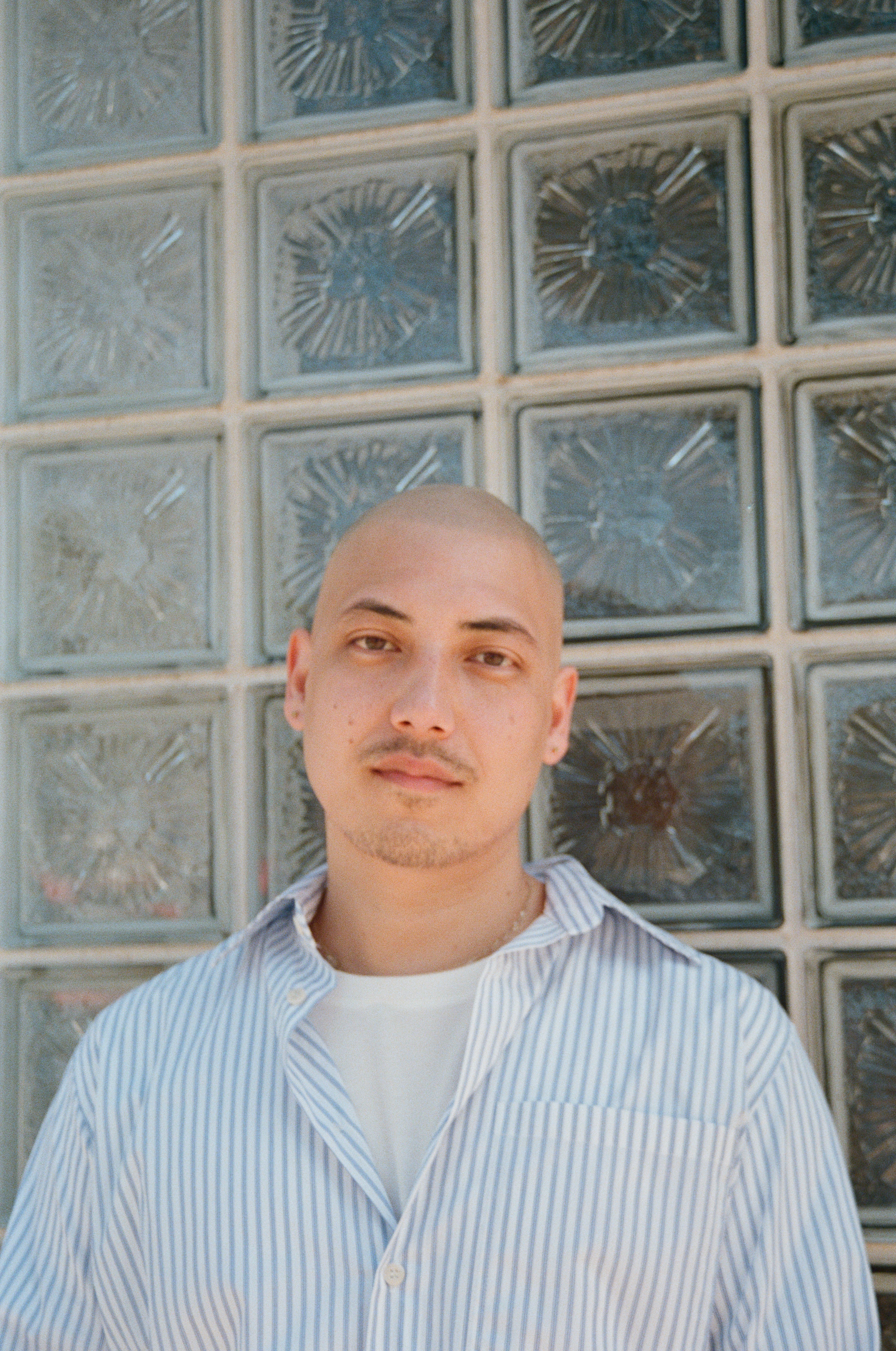 A photographic portrait of a man wearing a blue and white striped collared shirt with a shite t-shirt underneath stands in front of a glass wall