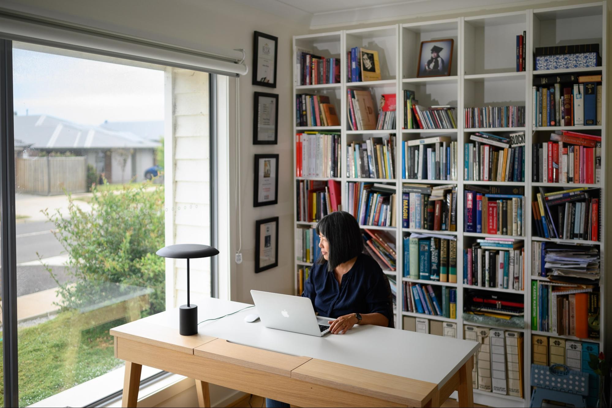 A wide shot of a woman sitting at a desk with her laptop, floor-to-ceiling bookshelves behind her. The woman is looking out the window.