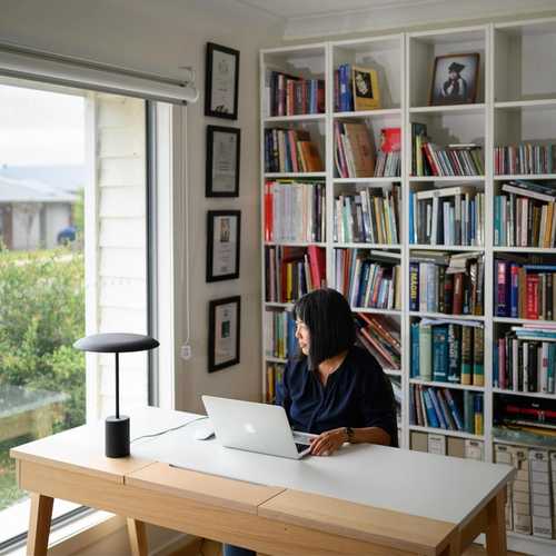 A wide shot of a woman sitting at a desk with her laptop, floor-to-ceiling bookshelves behind her. The woman is looking out the window.