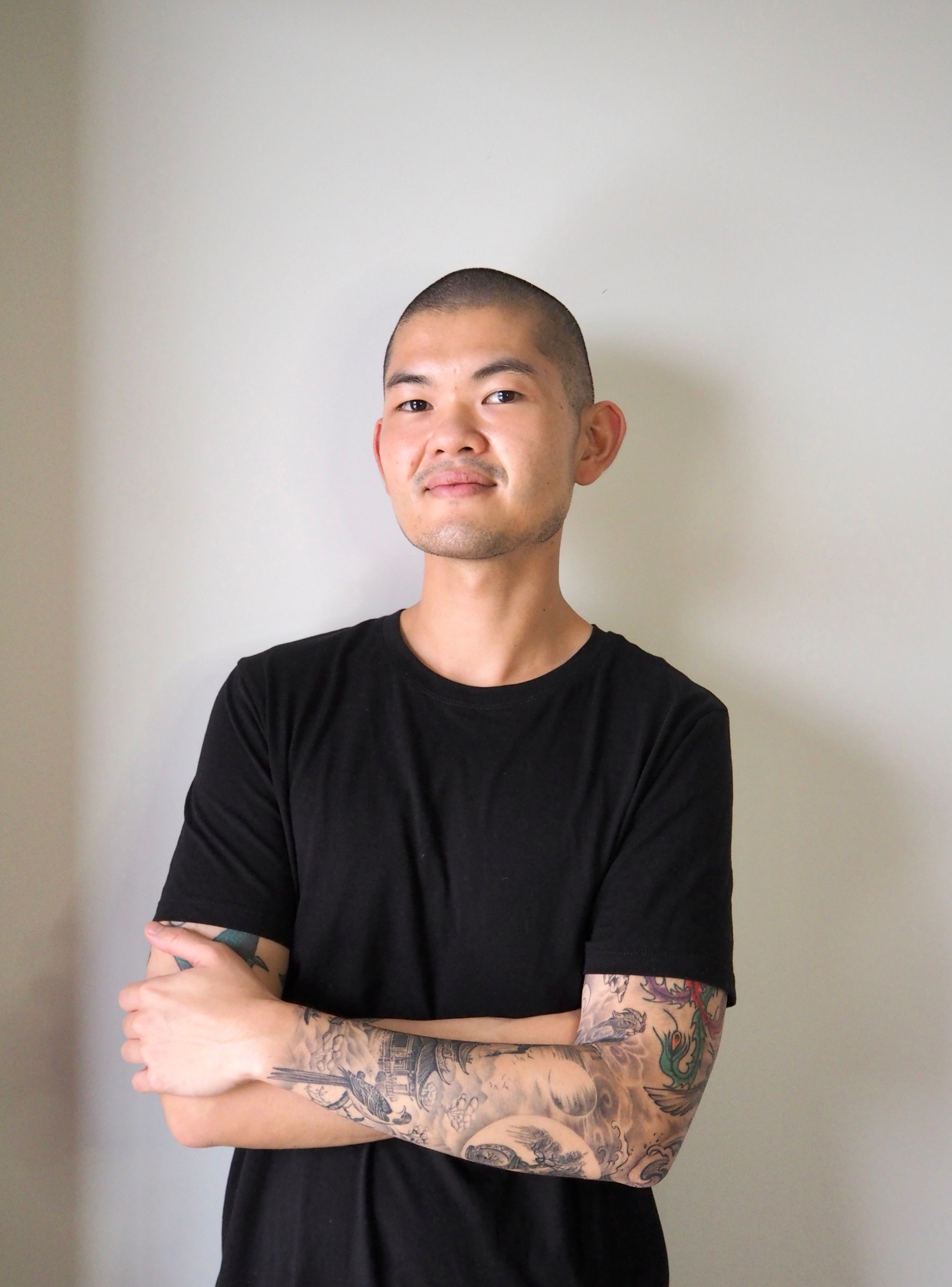 A man standing against a plain white wall, wearing a black t-shirt. He has tattoos on his left arm.