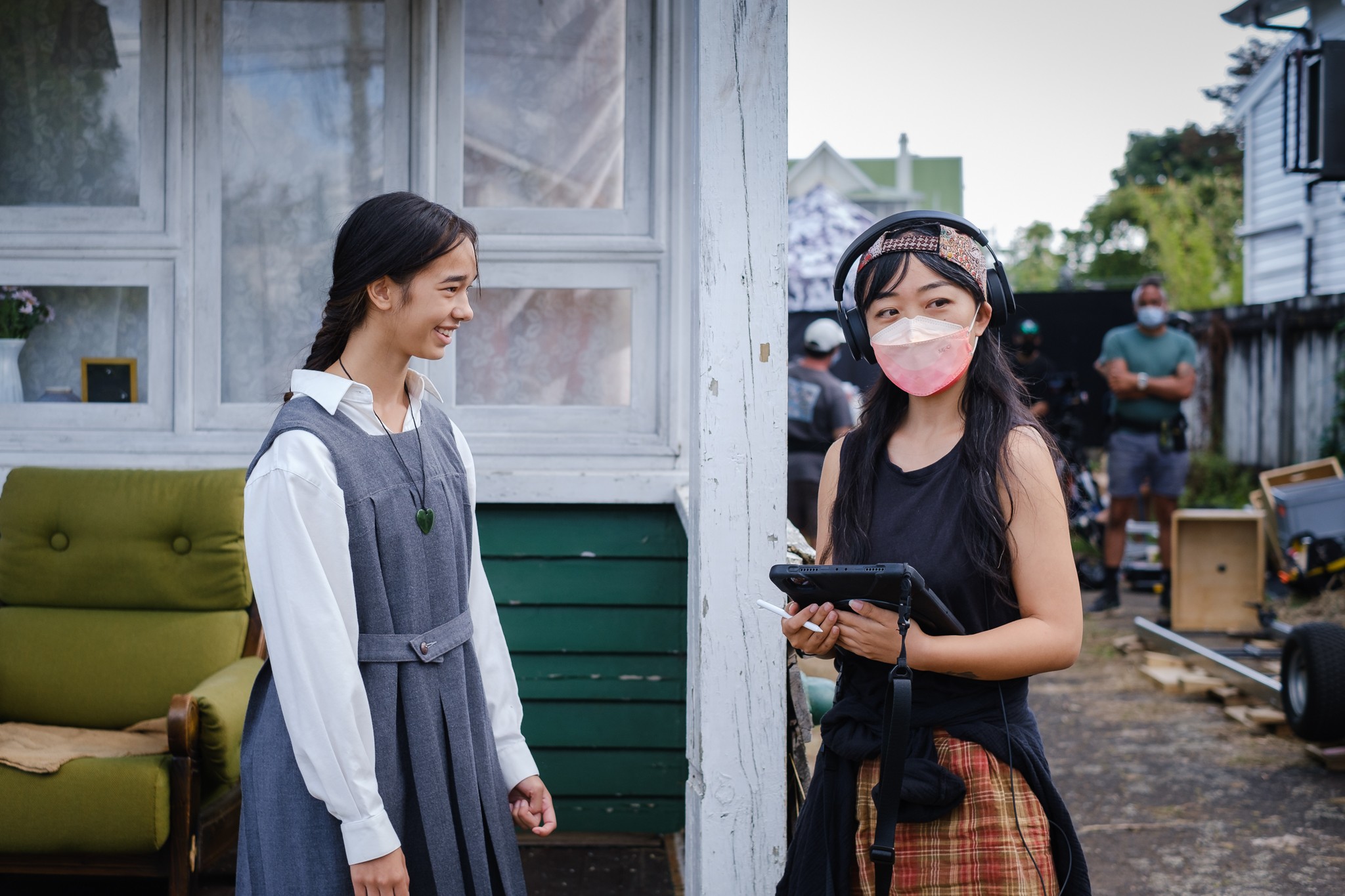 Two people standing outdoors on a film set. One is wearing headphones and holding an ipad while wearing an N95 mask while the other (actor) is smiling at them