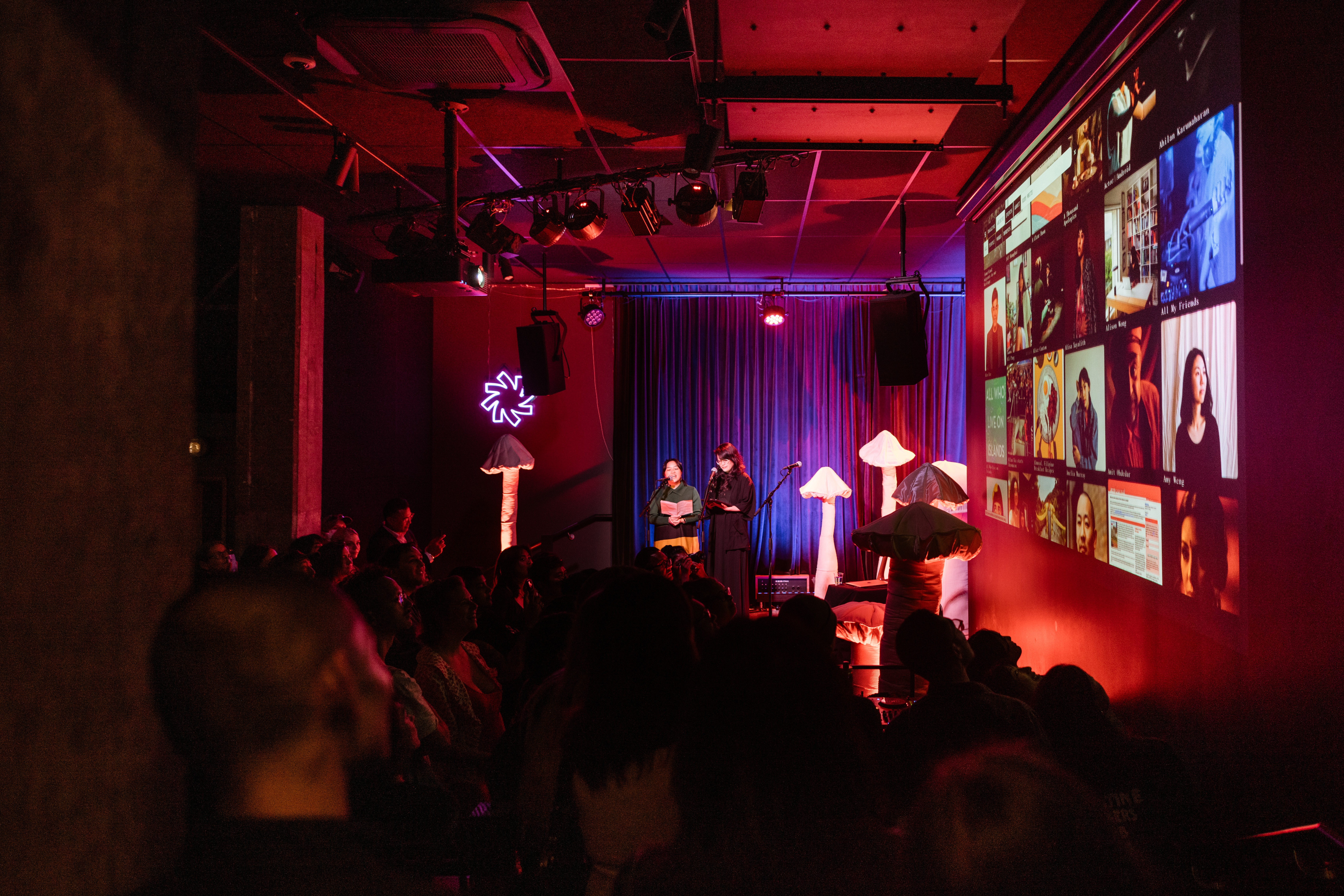 A crowd observing two people on stage surrounded by mushroom sculptures. A projection of a website is on the wall.