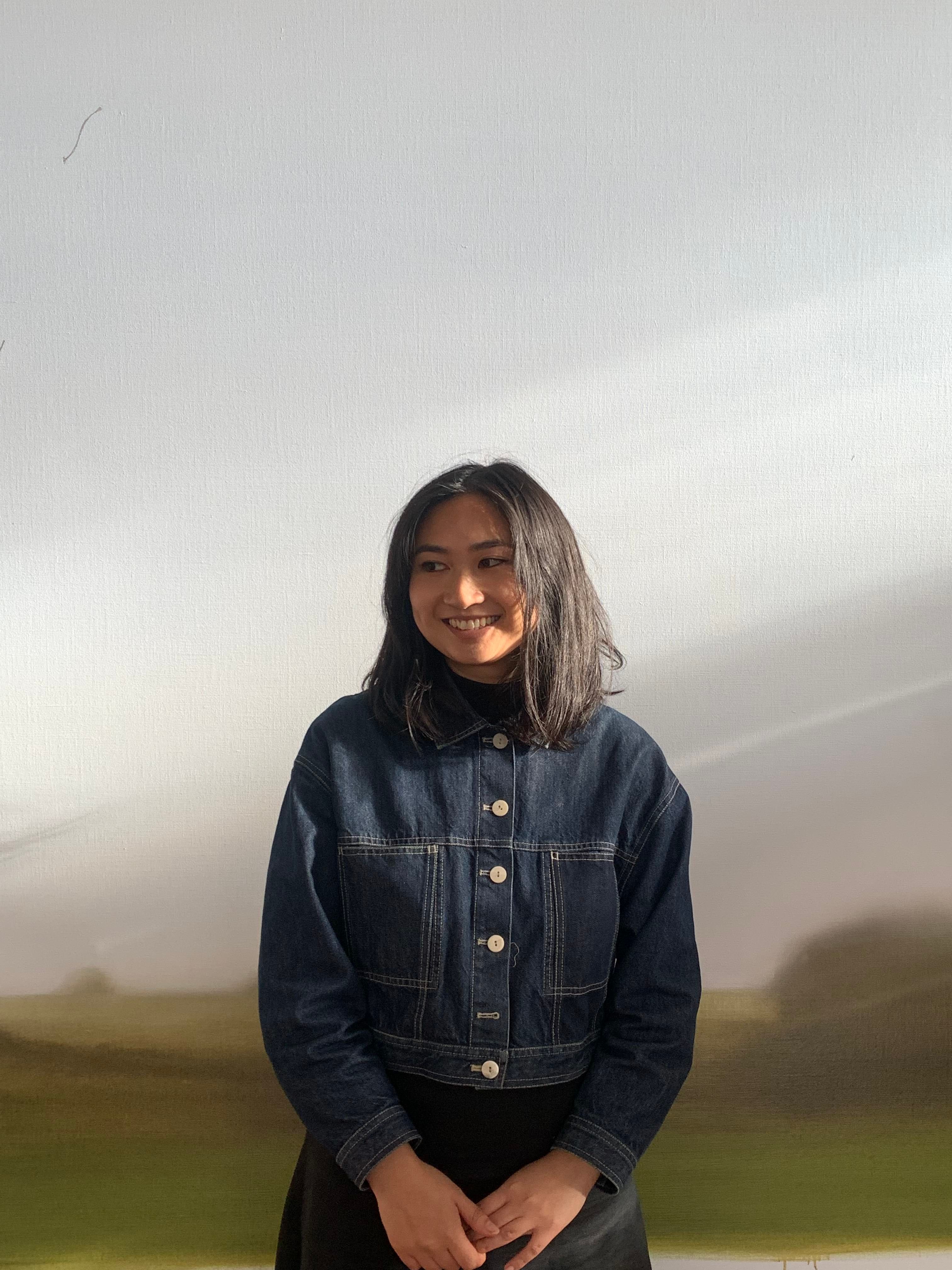 A woman with a black bob and a denim jacket stands in front of a wall