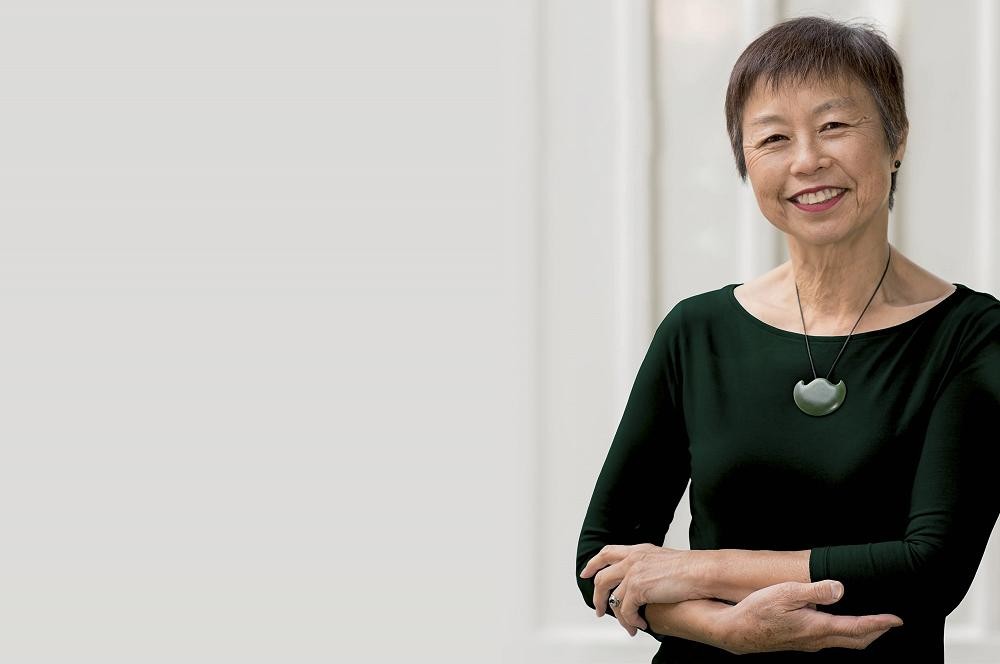 A woman with cropped black hair and a black blouse leans against a wall, smiling.