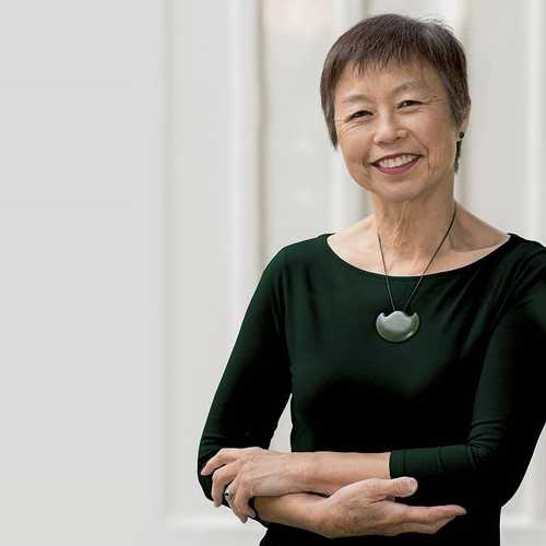 A woman with cropped black hair and a black blouse leans against a wall, smiling.