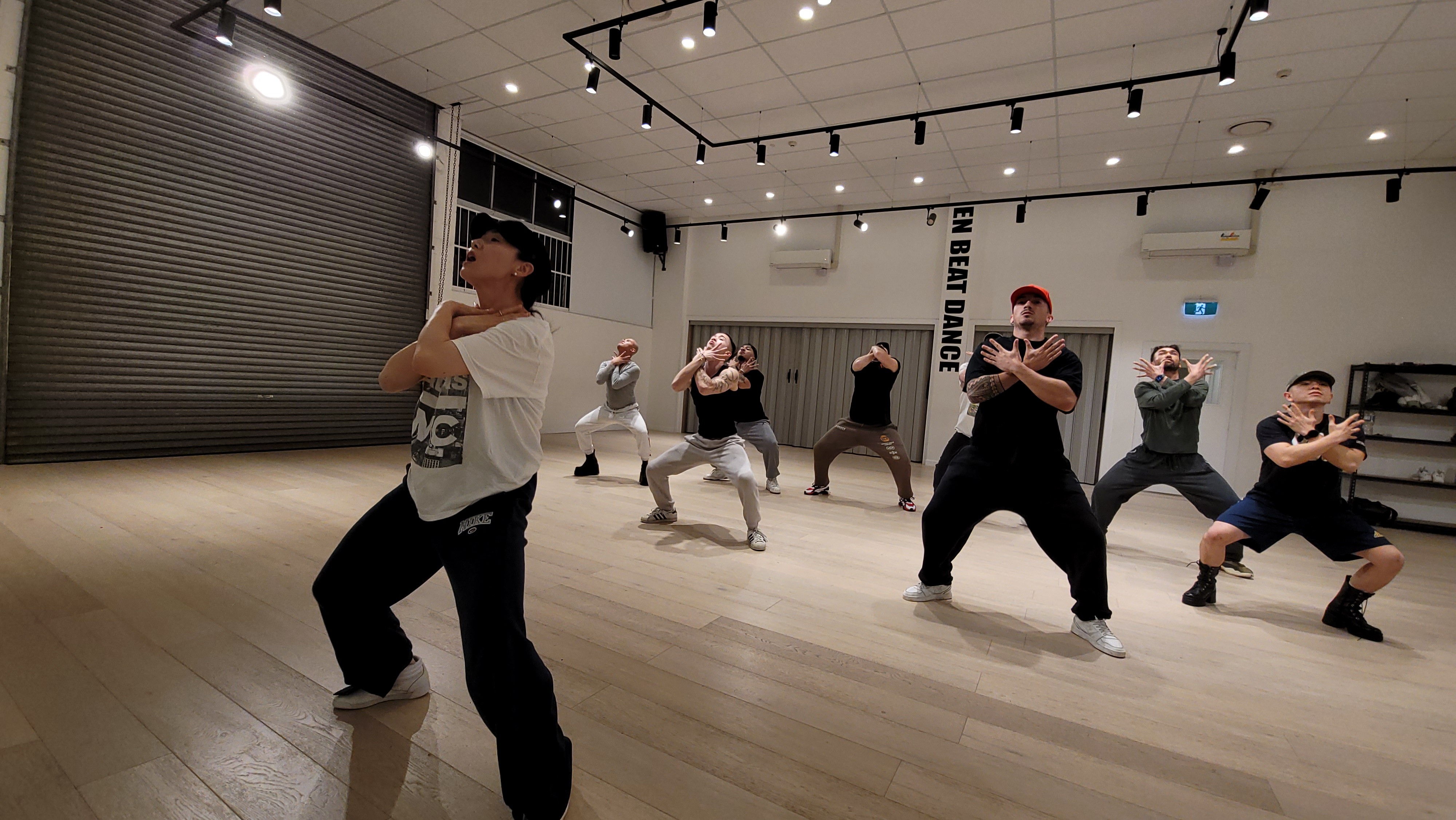 A woman leading a dance class with her arms crossed across her chest. She is wearing a white shirt, black pants and a cap