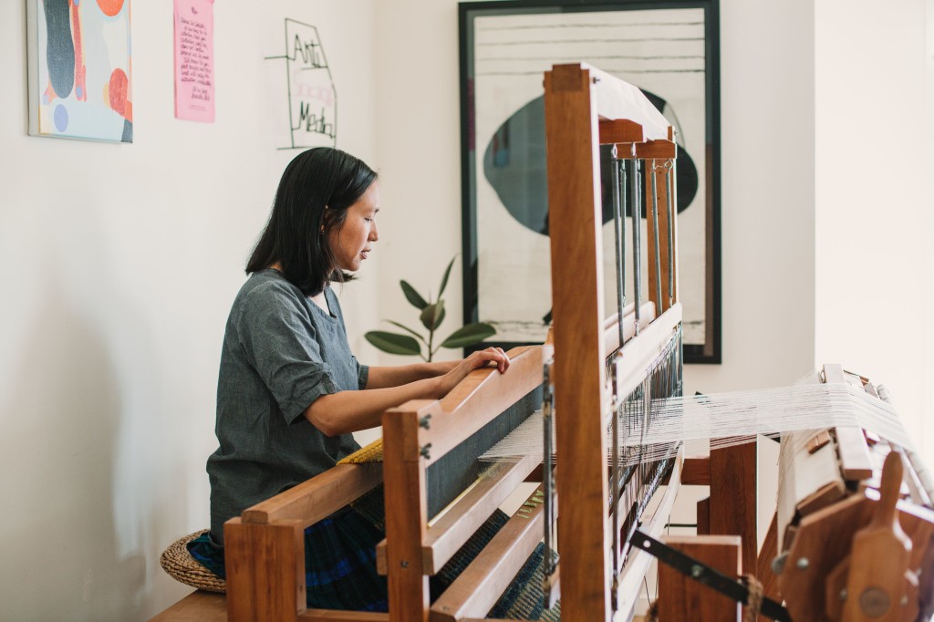 A woman sits at a weaving loom in a room hung with artwork