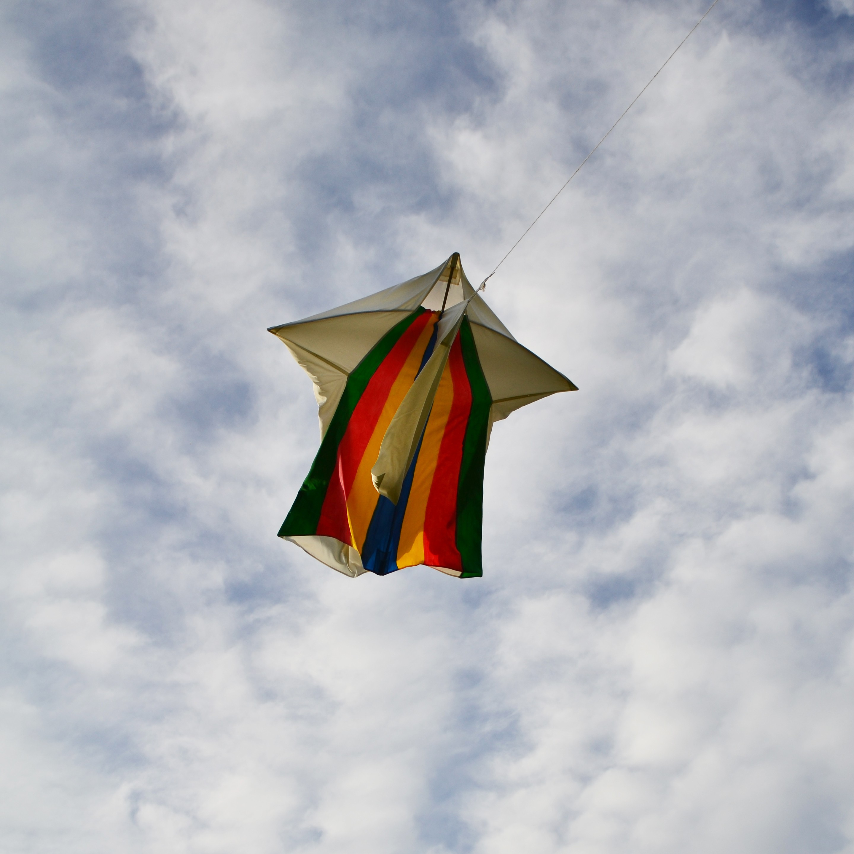 A kite with white and rainbox panels flying high against a cloudy sky.