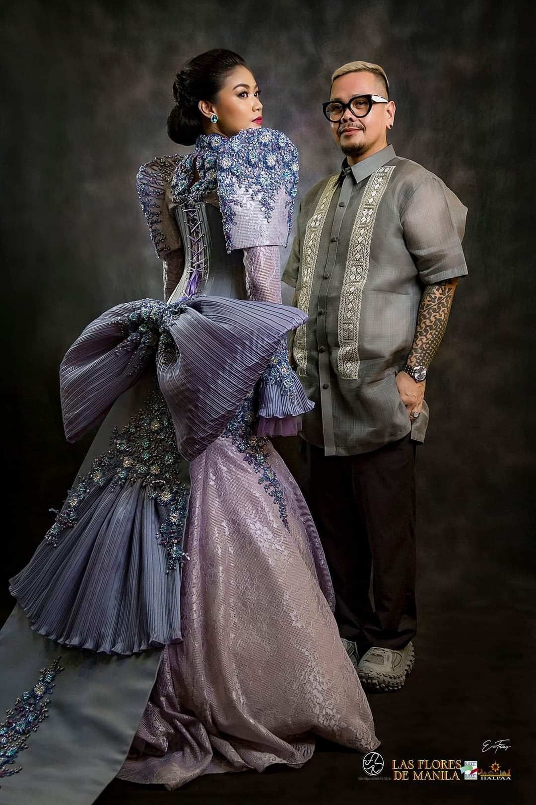 2 Filipinos standing side-by-side: one wearing an intricately beaded and constructed purple dress with their hair in a tight bun, the other wearing a black horn rimmed glasses, translucent grey embroidered top, black pants and grey sneakers