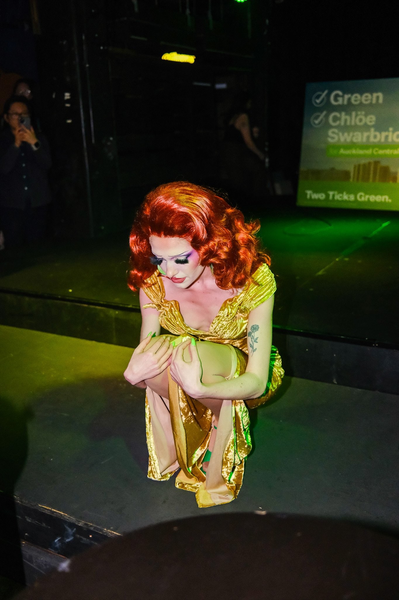 A drag queen wearing a 50s style red wig and gold outfit posing dramatically while performing on stage