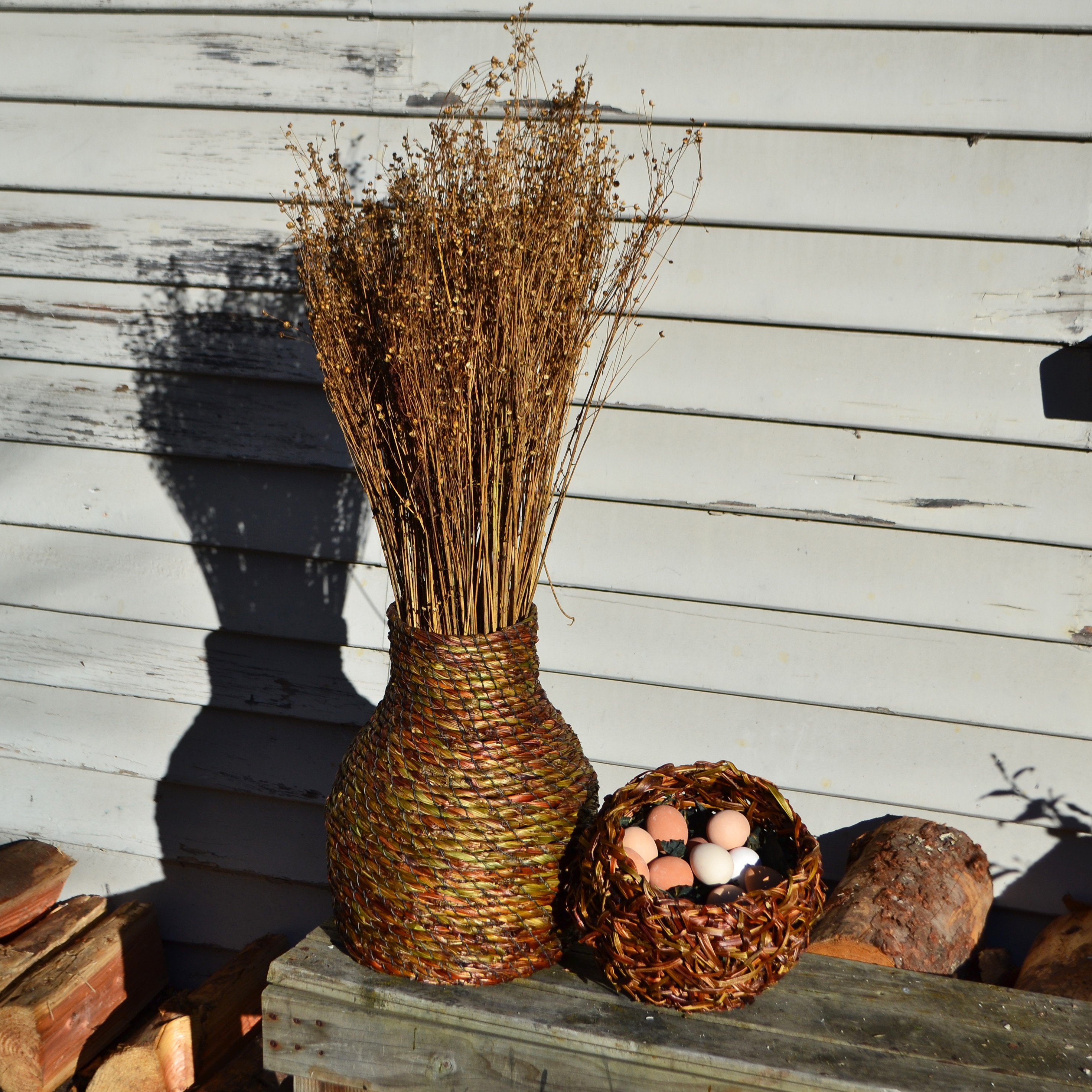 A vase and basket made of leaves and stems, holding more stems and ceramic eggs.