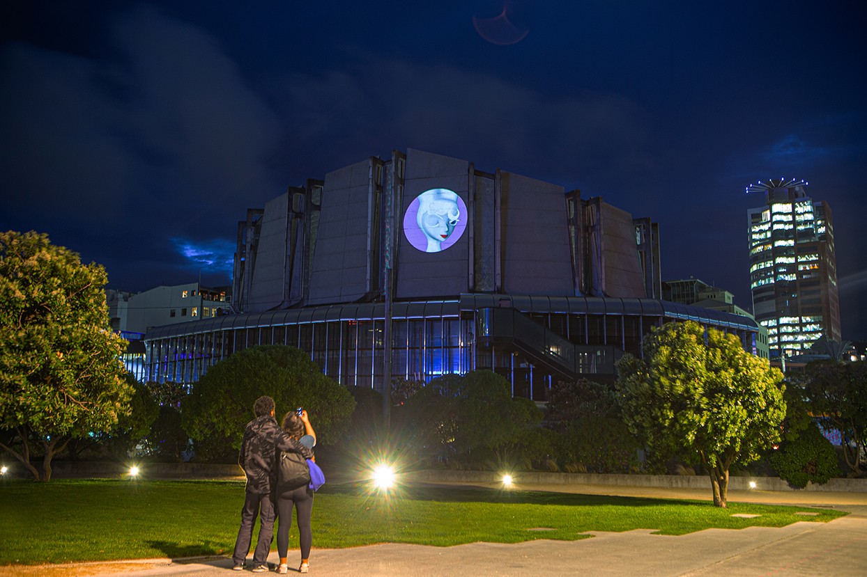 A circular projection on a brutalist building shows the head of an animated bunny-woman character.