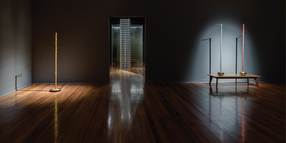 Spot lights on two sculptures depicting noodles being pulled from bowls by chopsticks to a greater than human hight, and a tall stack of stainless steel crockery. 