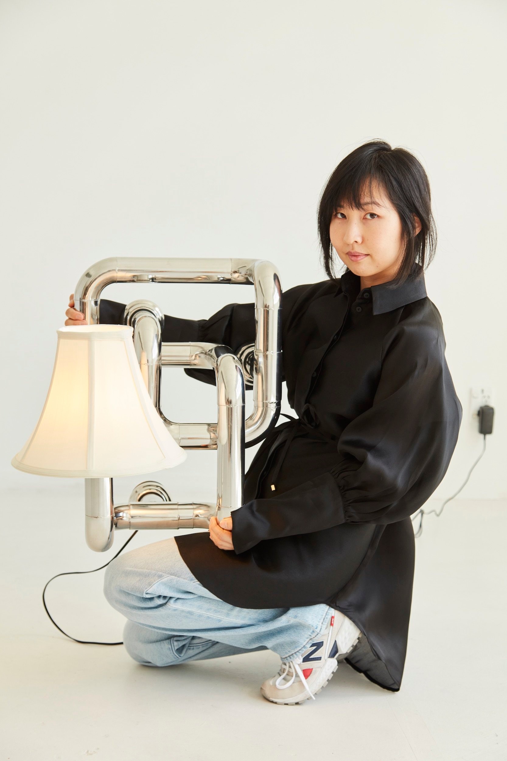 A woman with black hair kneels while holding a sculpture bent from shiny steel pipe with an illuminated lamp shade