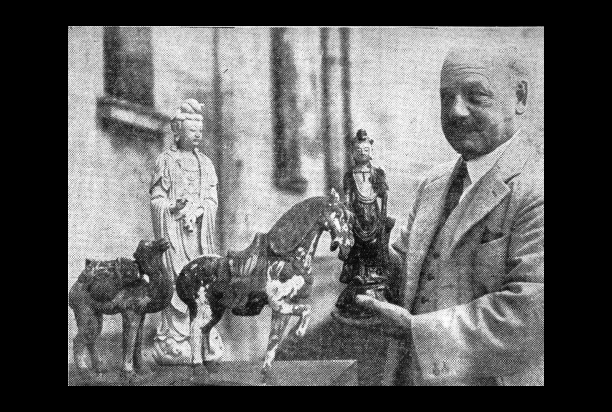 A man in a three-piece suits stands holding Chinese artworks.