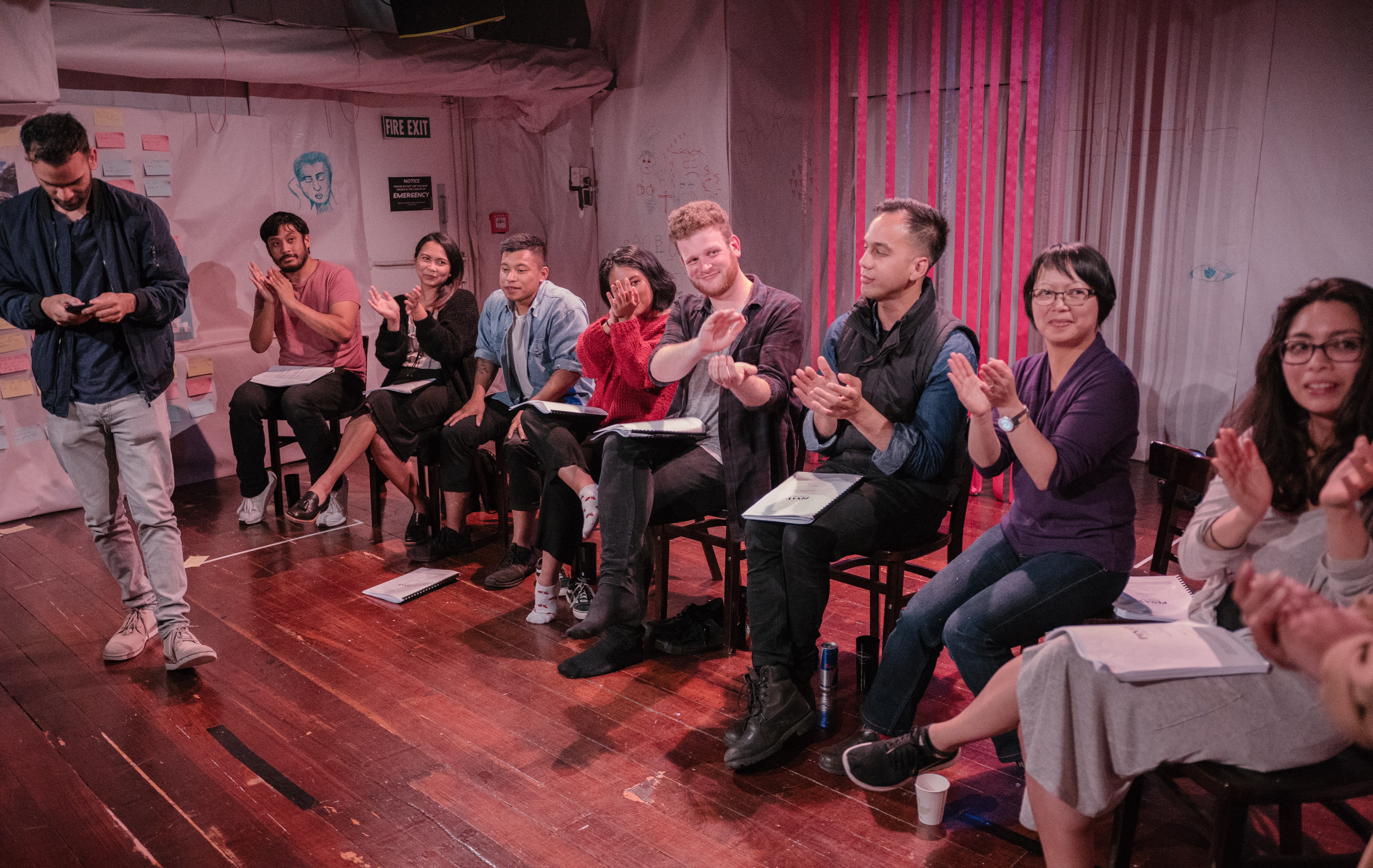 Nine people sit on chairs with scripts in their lap, clapping towards someone offstage