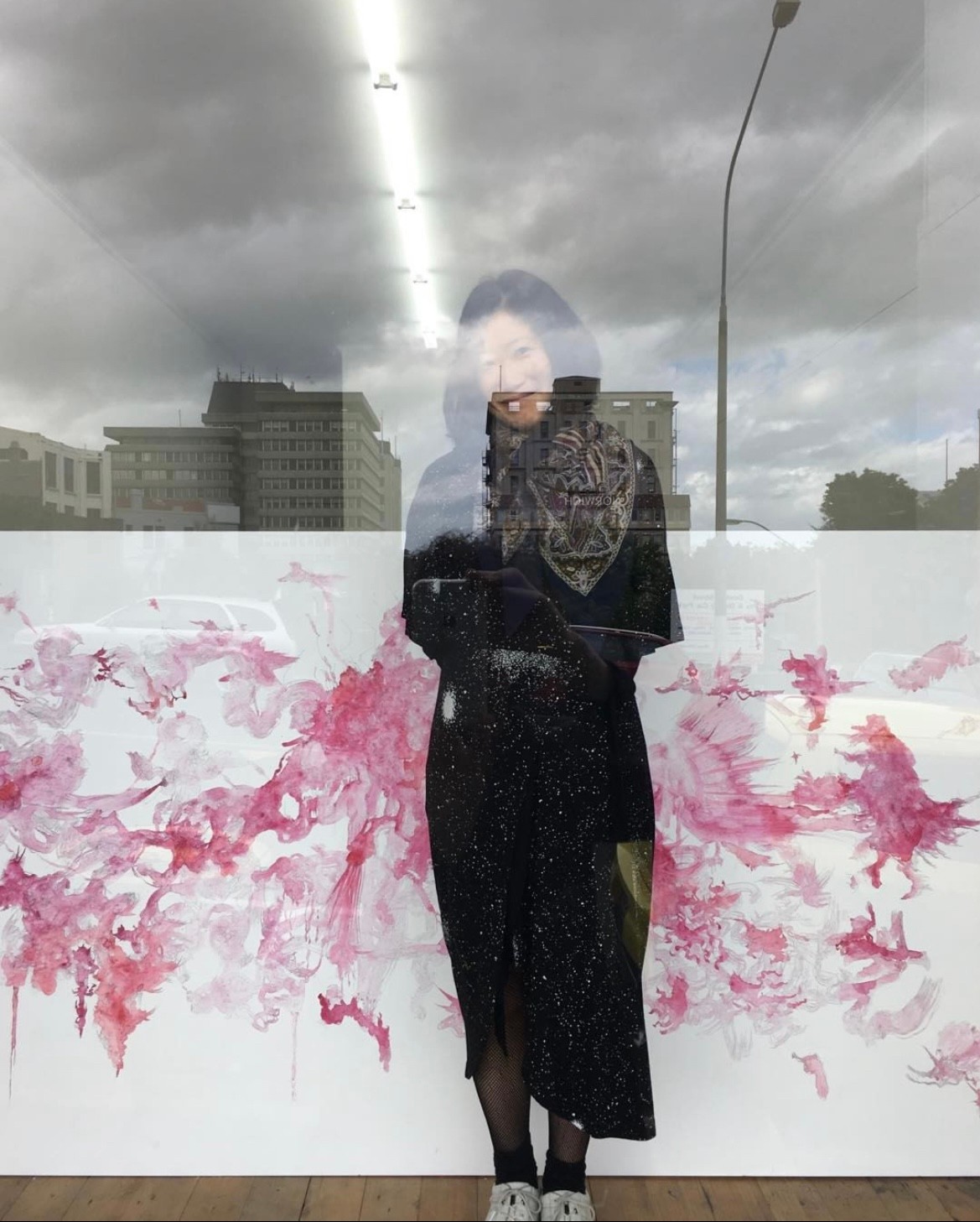 Dark haired woman wearing a long black dress standing in front of a painting and looking out from behind reflective glass.