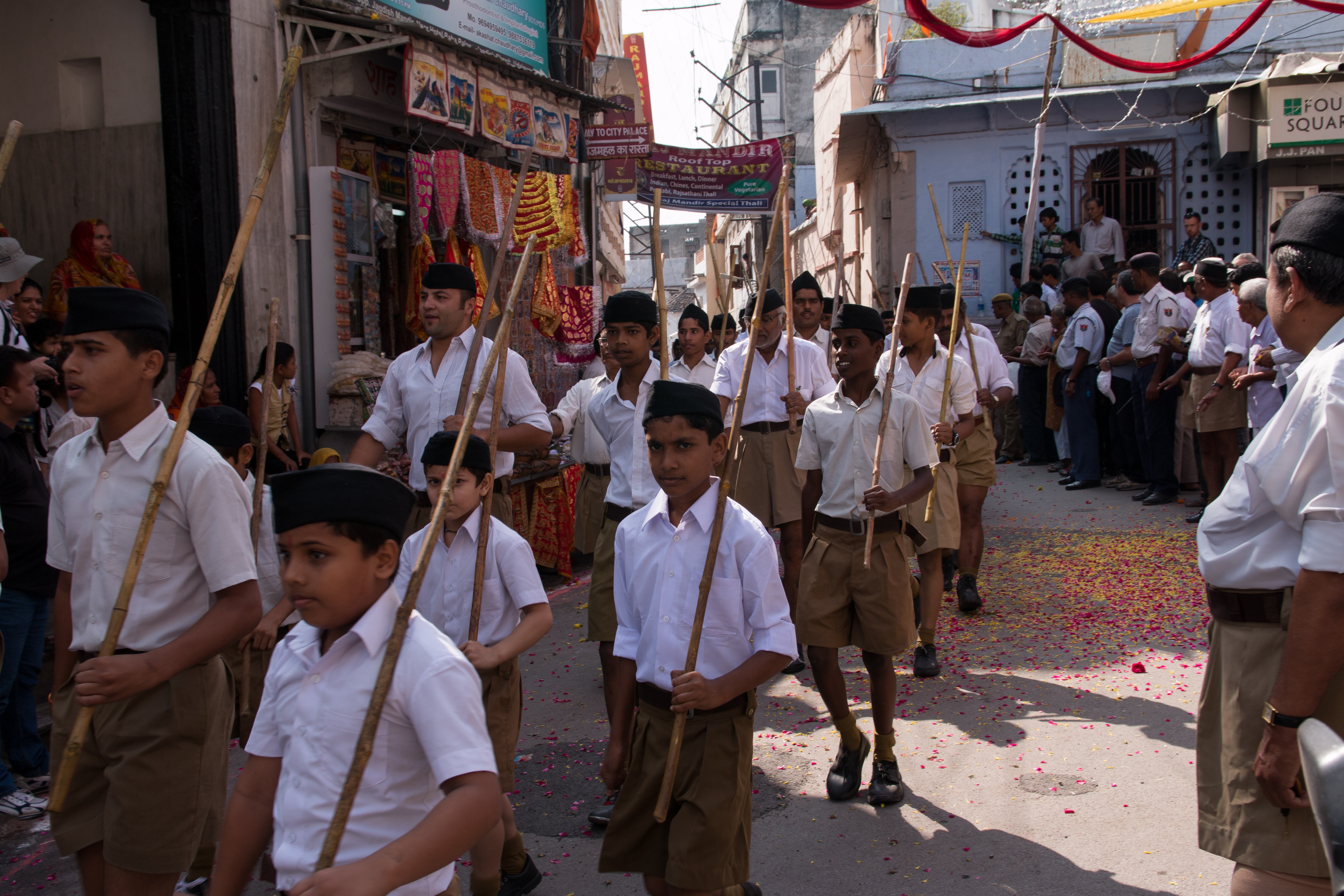 Children with lathi (long sticks) and wearing uniforms parade through a street.