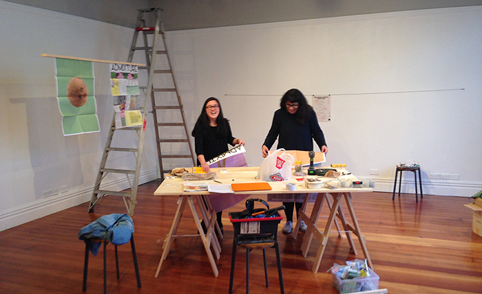 A Chinese woman and a Samoan women stand behind a trestle table in an art gallery while an exhibition is being set up.